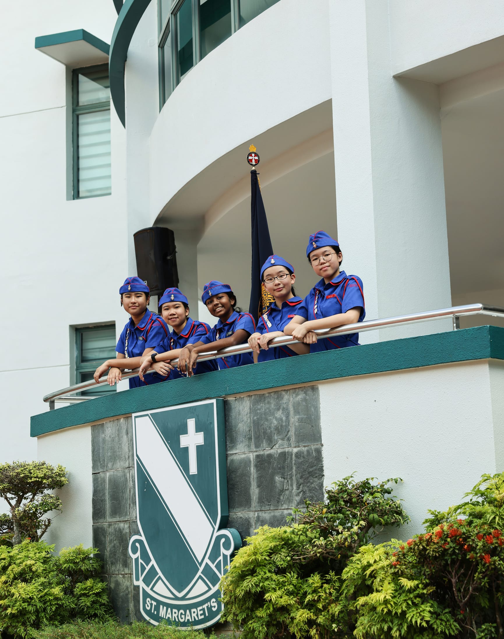 Five girls in blue St. Margaret's school uniforms stand on a balcony above the school's crest, with a flag behind them.