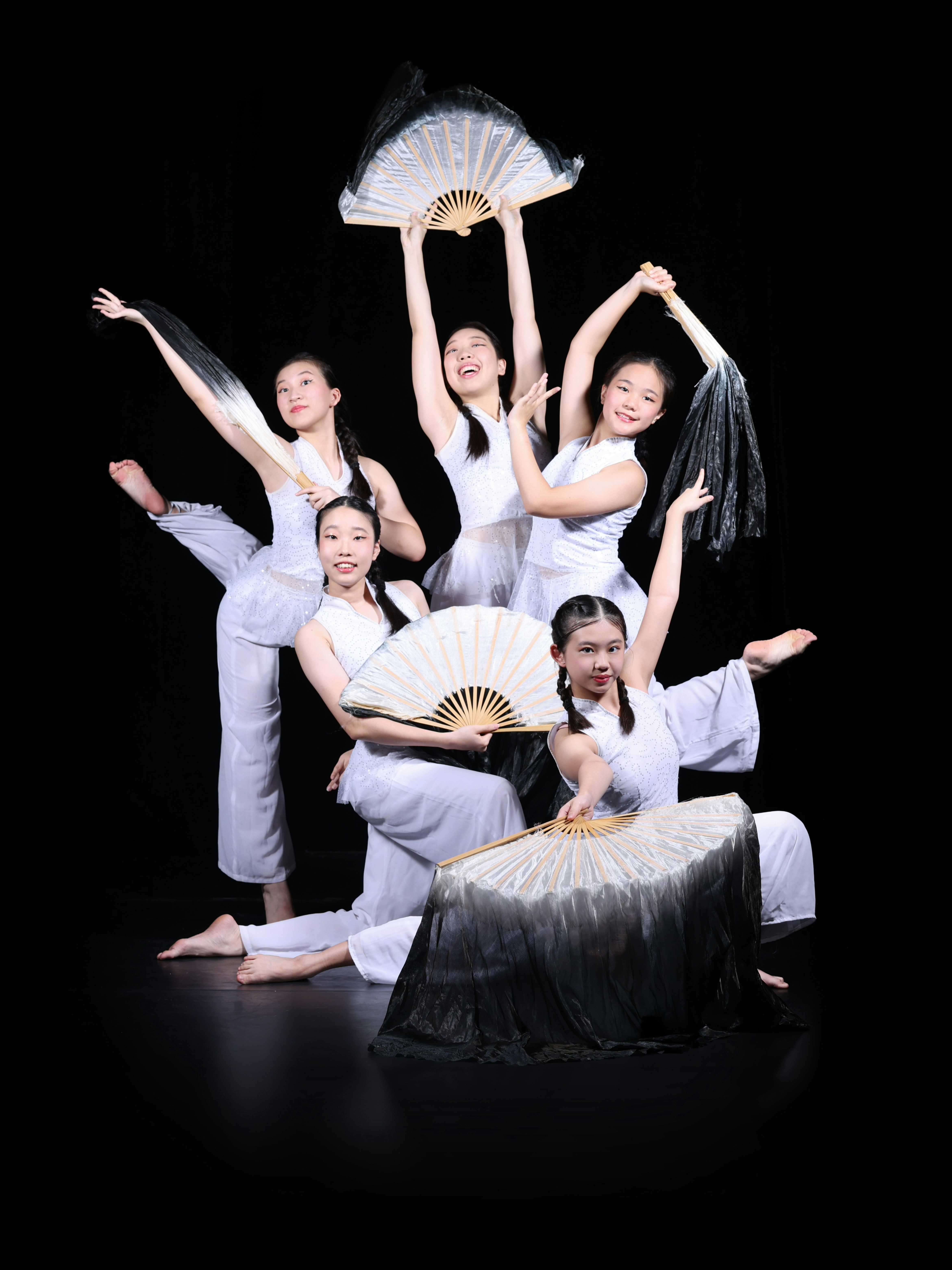 Five young girls in white costumes pose gracefully holding large Chinese fans against a black background.