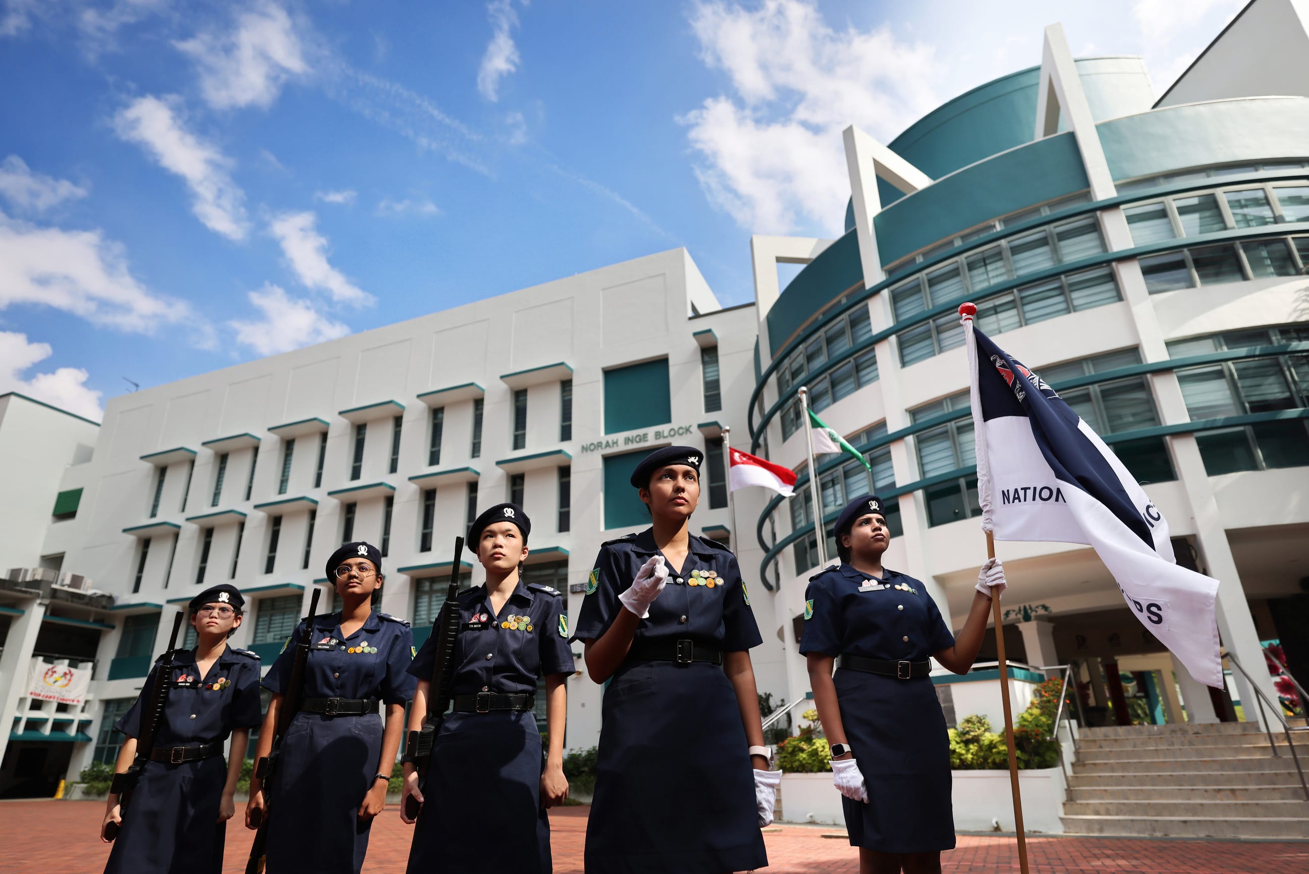 Five female cadets in navy uniforms stand at attention outside Norah Inge Block, one holding a flag.