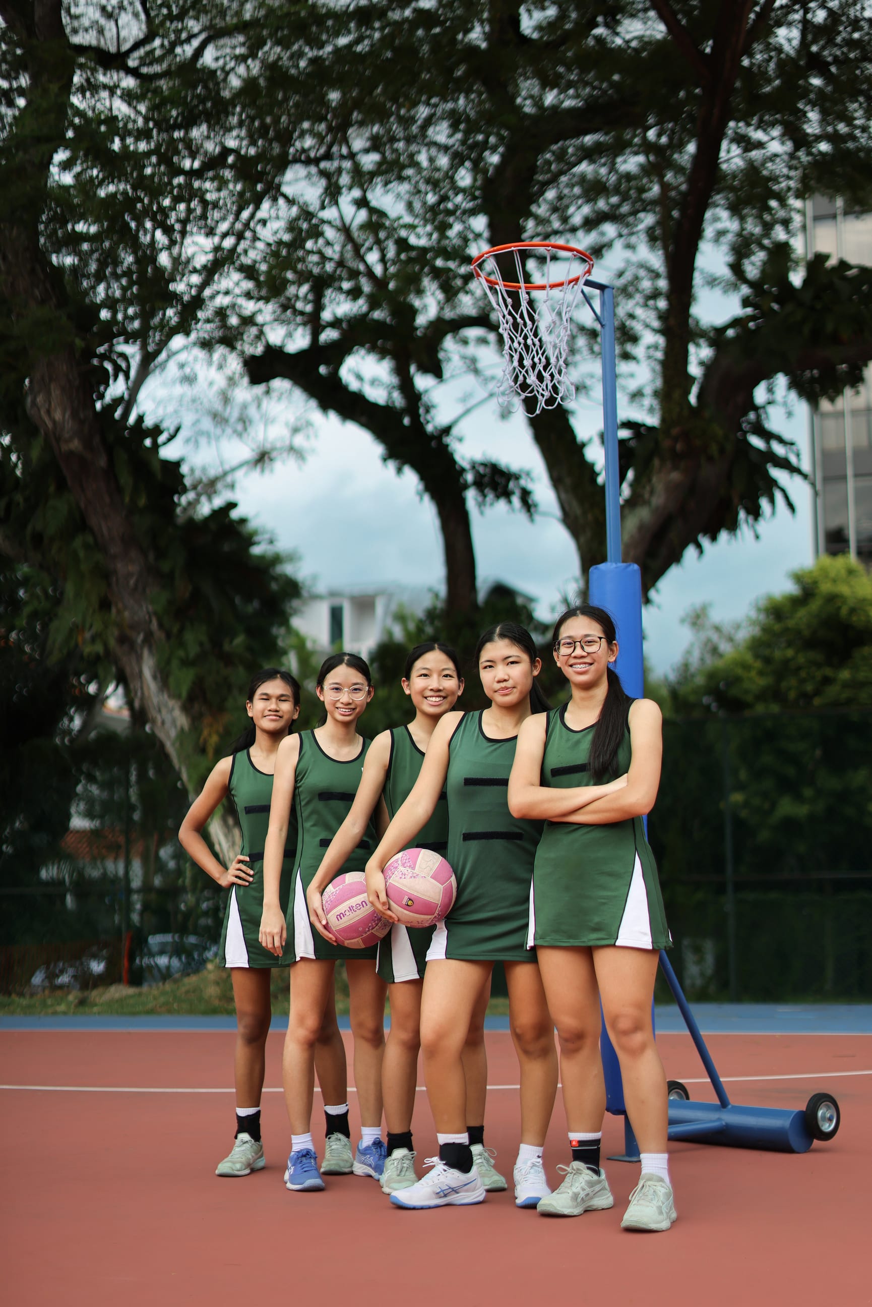 Five players in green uniforms pose on an outdoor court by a hoop; two hold pink basketballs.