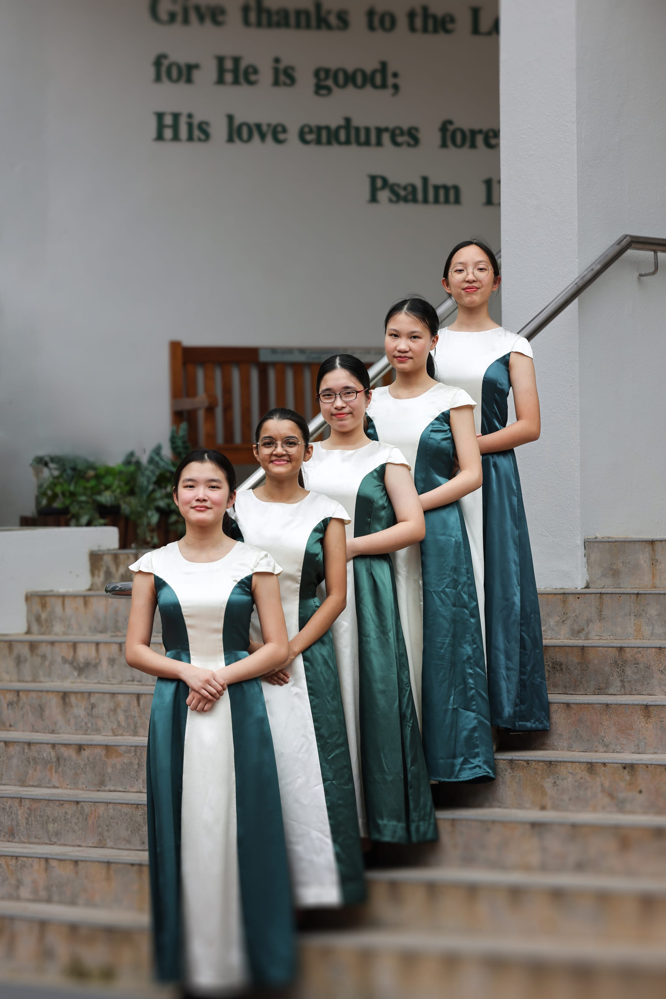 Five girls in matching teal-and-white dresses stand on stairs beneath a wall with a Bible verse.