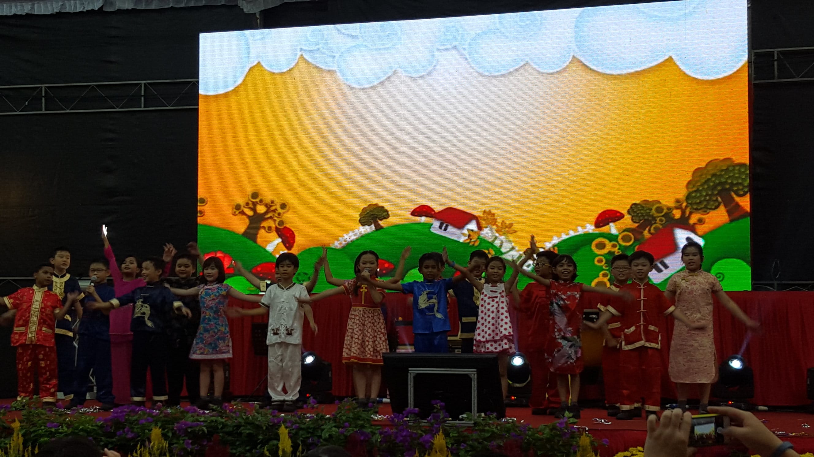 Children in traditional Chinese costumes perform on a stage with a colorful cartoon landscape on a large LED screen behind them.