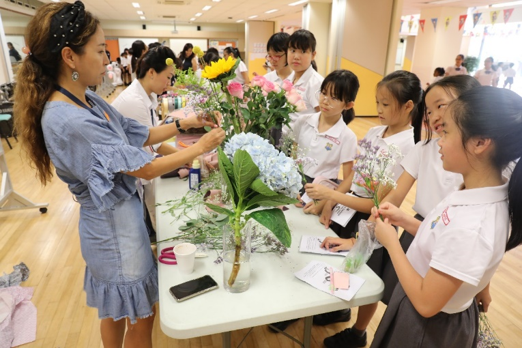 A woman in a blue dress teaches children flower arranging at a table with colorful blooms including hydrangeas.