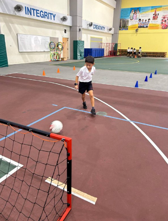 A young boy in school uniform kicking a soccer ball toward a small goal on an indoor sports court.