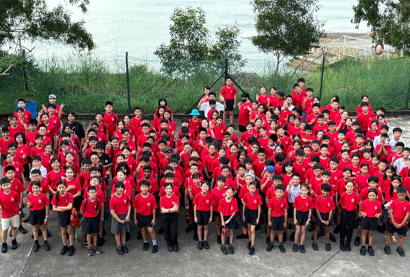 Large group of students in red uniforms posing outdoors near a waterfront with green trees behind them.
