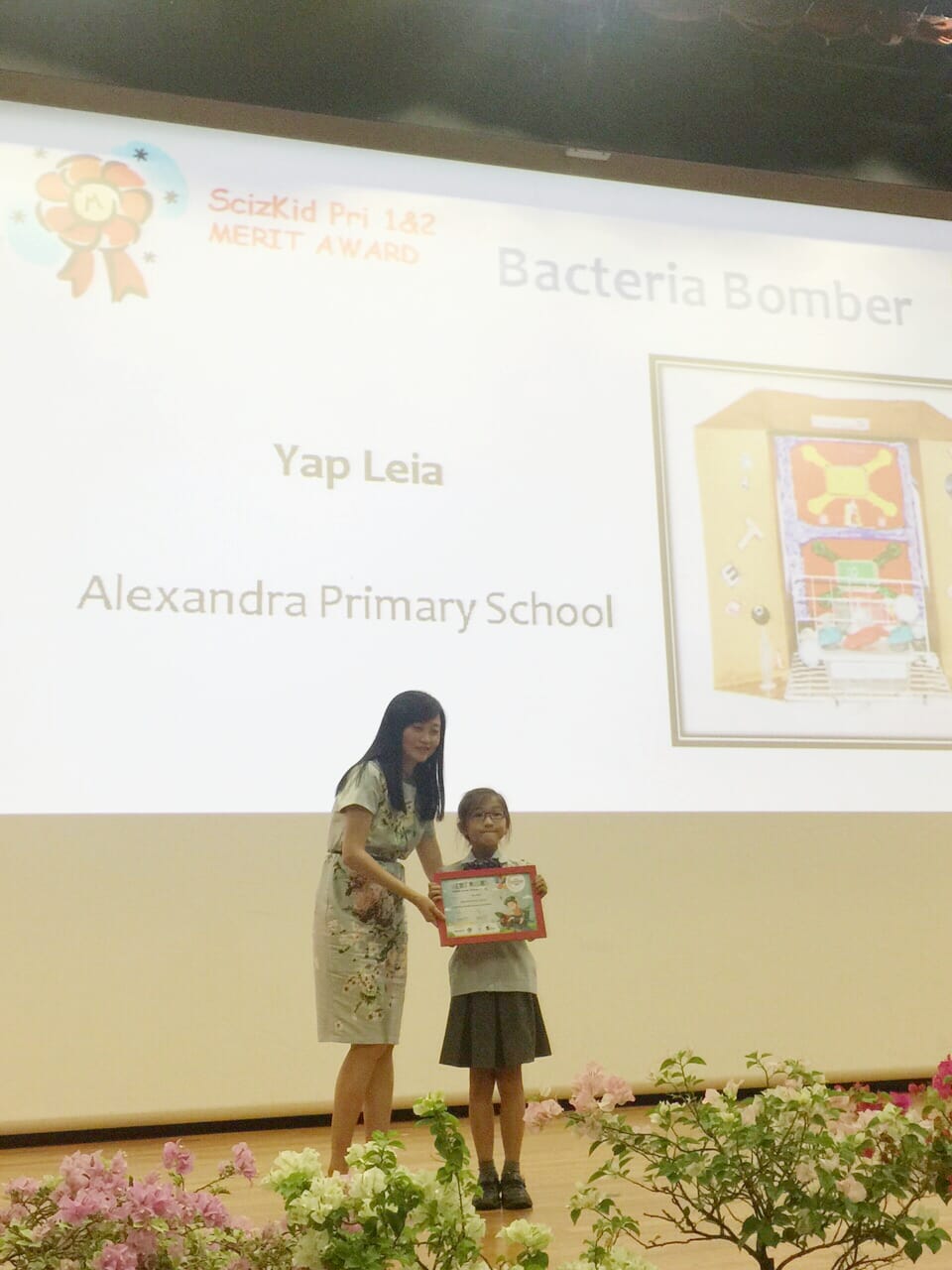 A woman presents a framed certificate to a young girl on stage at a ScizKid Merit Award ceremony.