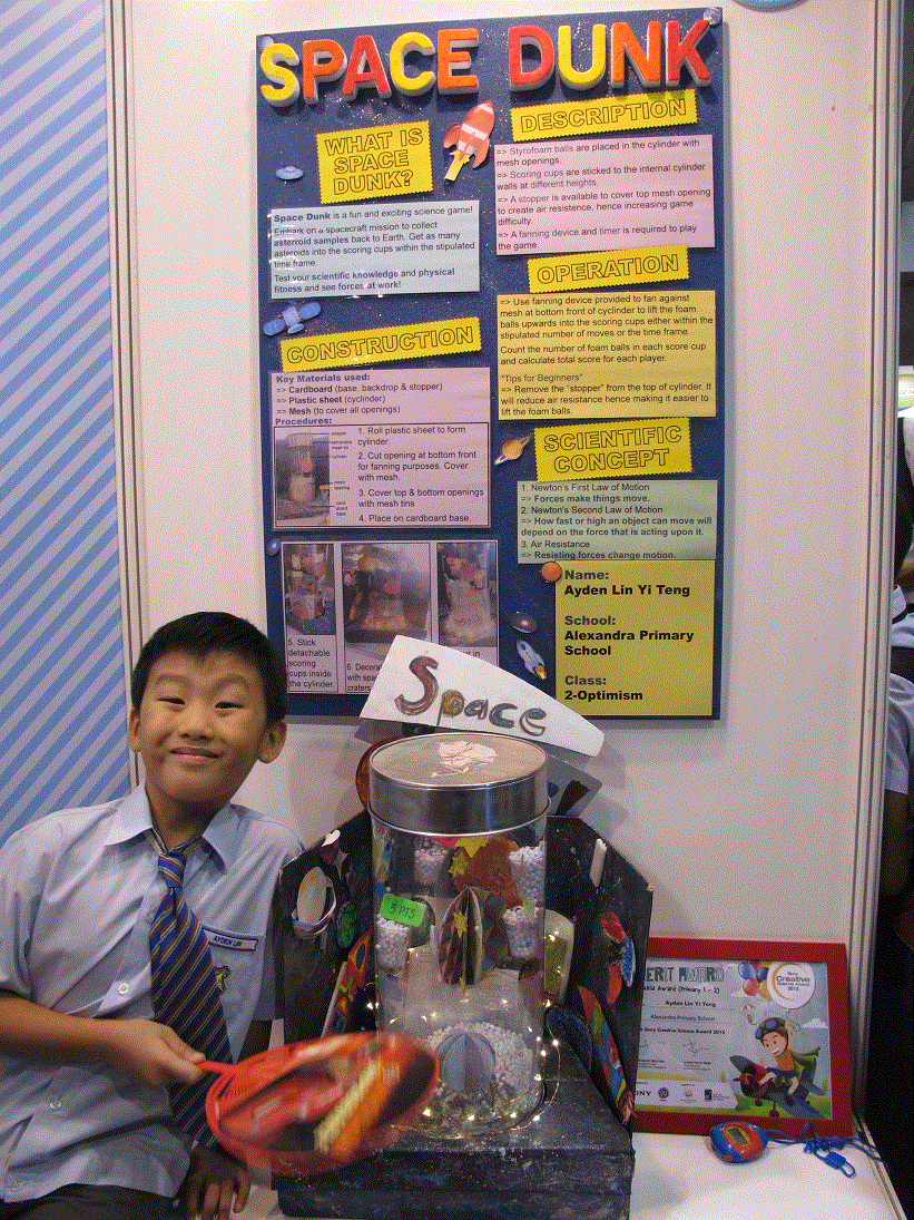 Boy in school uniform sits beside a science fair project titled Space Dunk with a poster and cylindrical model.