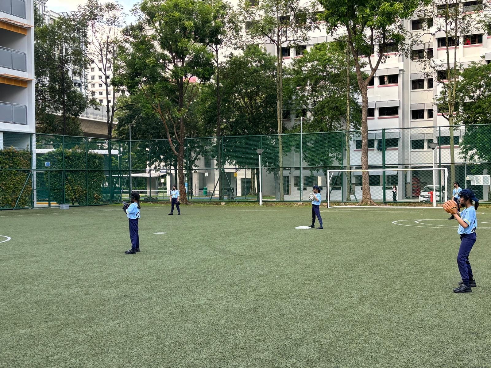 Students in blue uniforms practice baseball on a green turf field surrounded by a fence and buildings.