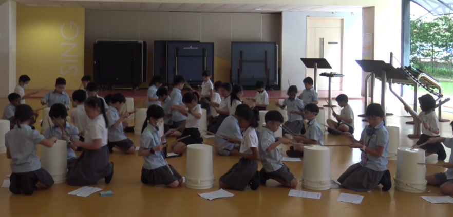 Children in school uniforms kneel on a floor, each drumming on a white bucket in a bright indoor space.