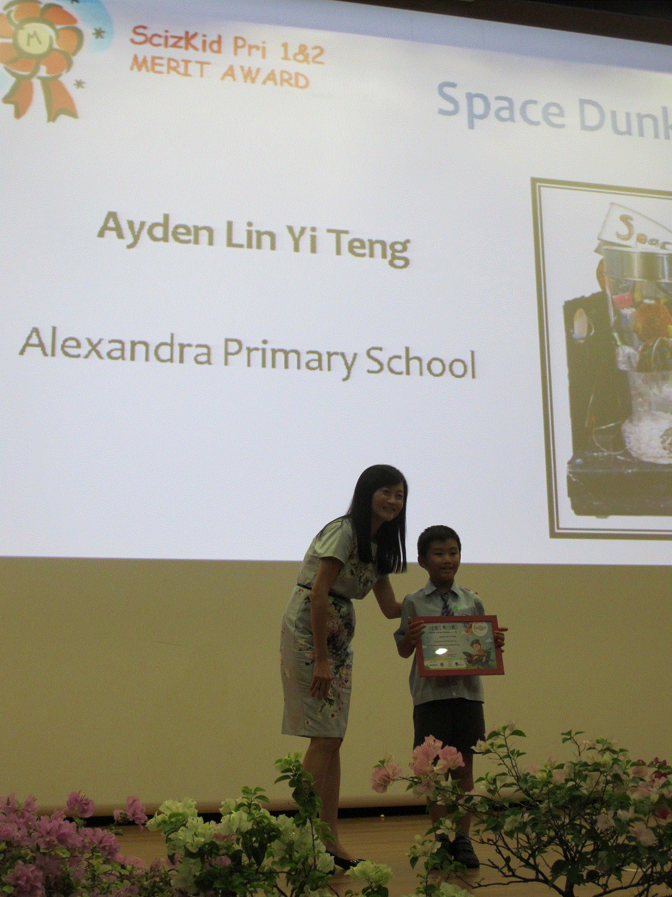 A woman and a young boy hold a certificate on stage at a ScizKid Merit Award ceremony.