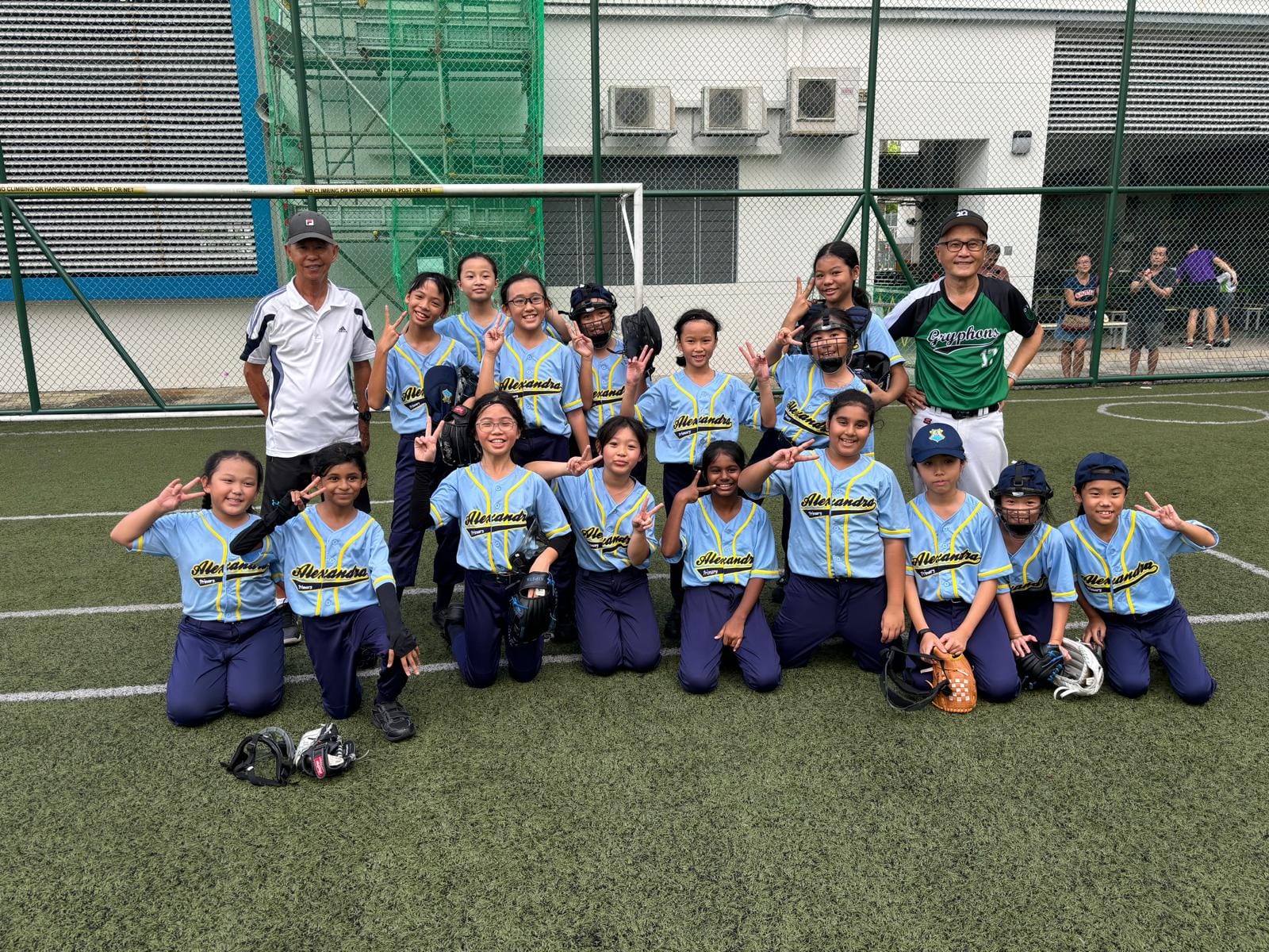 A youth girls softball team in light blue Alexandra jerseys poses with two coaches on a green turf field.