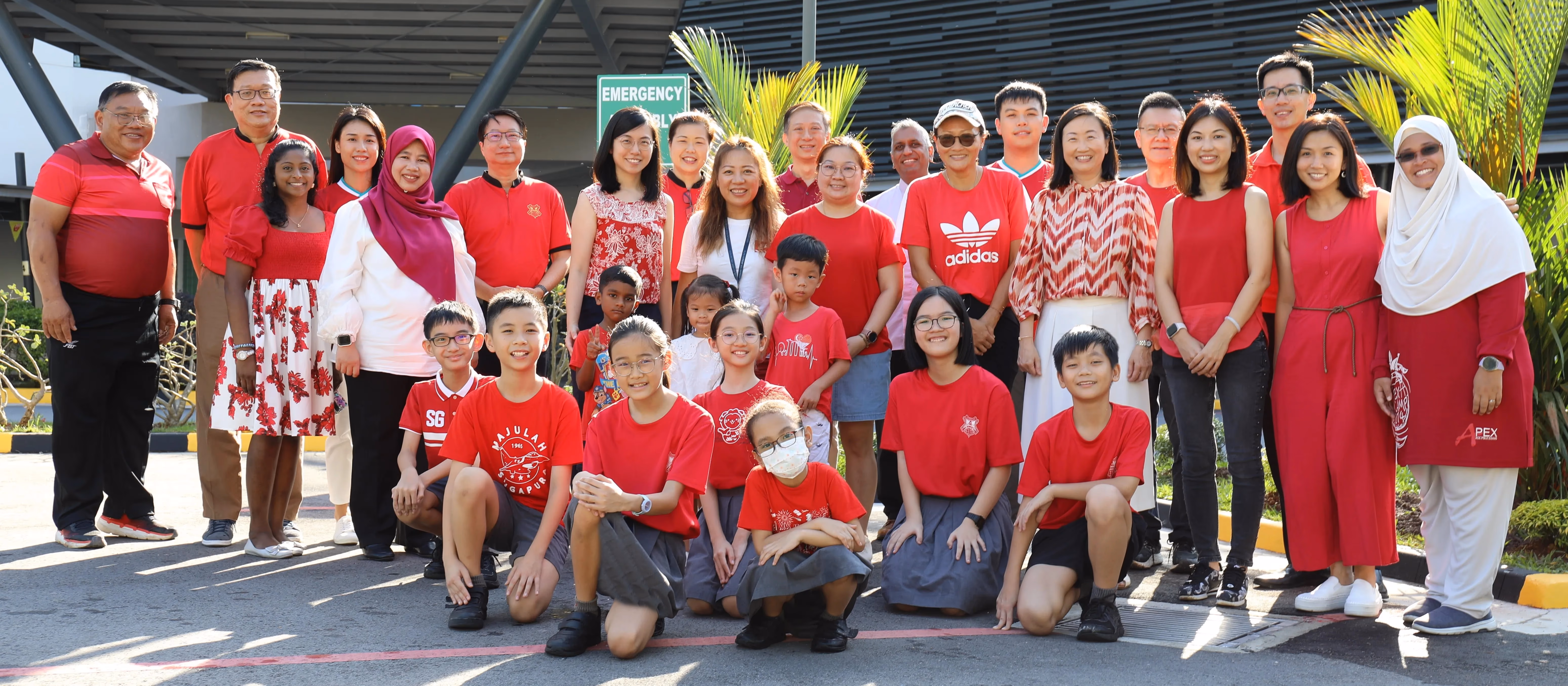 A large group of adults and children wearing red outfits pose together outdoors near an emergency assembly point sign.