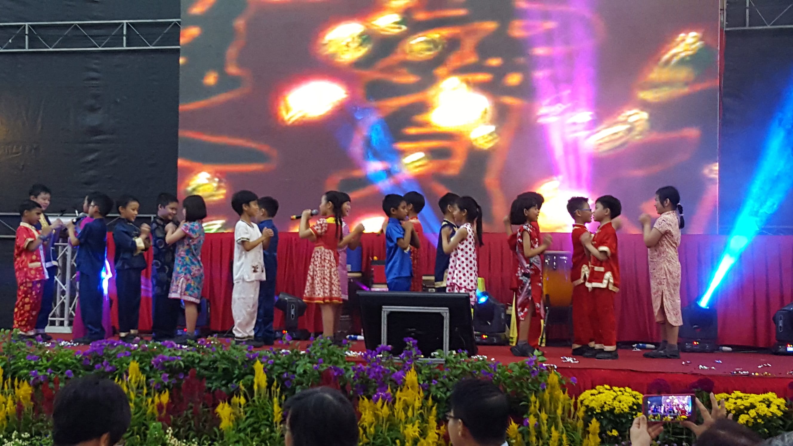 Children in colorful traditional and casual outfits perform on a decorated stage with bright lights and a large screen behind them.