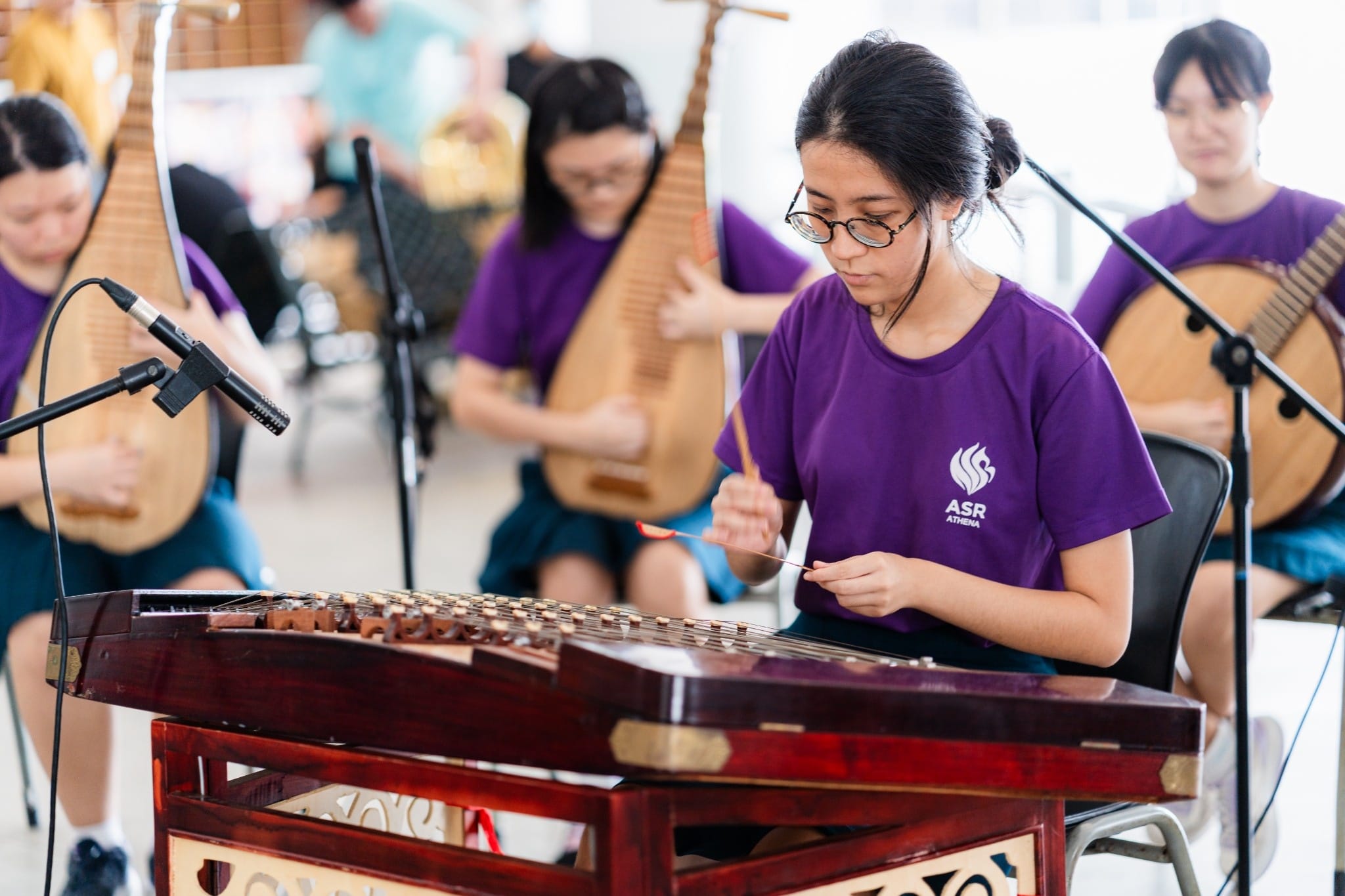 Young musicians in purple ASR Athena shirts play traditional Chinese instruments including a yangqin and pipa.