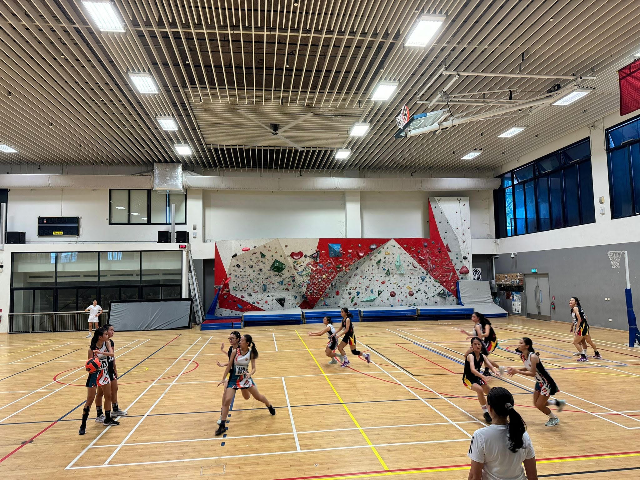 Girls playing basketball in an indoor court with a colorful climbing wall in the background.