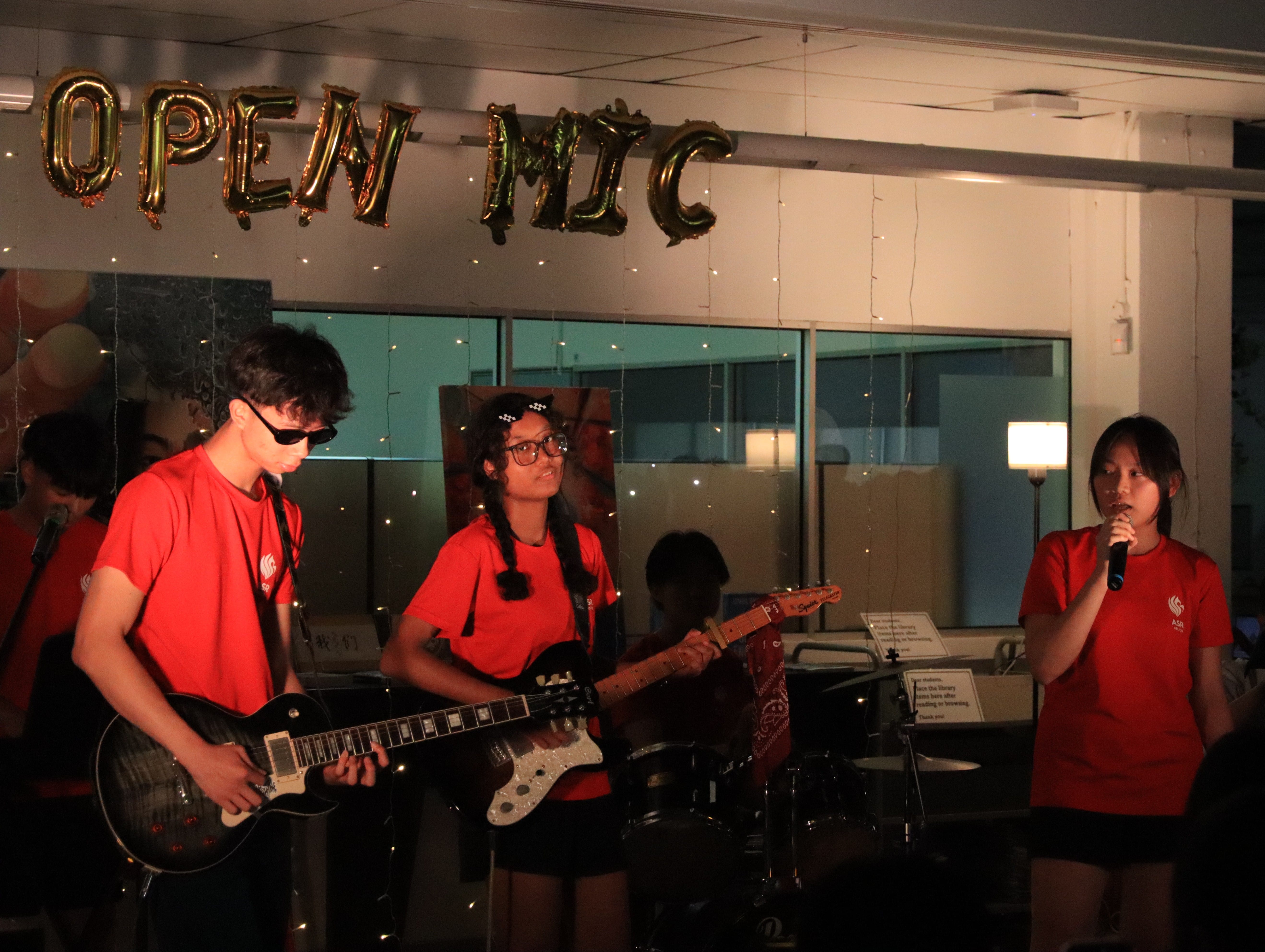 Three teens in red shirts perform at an open mic event, two playing guitars and one singing into a microphone.
