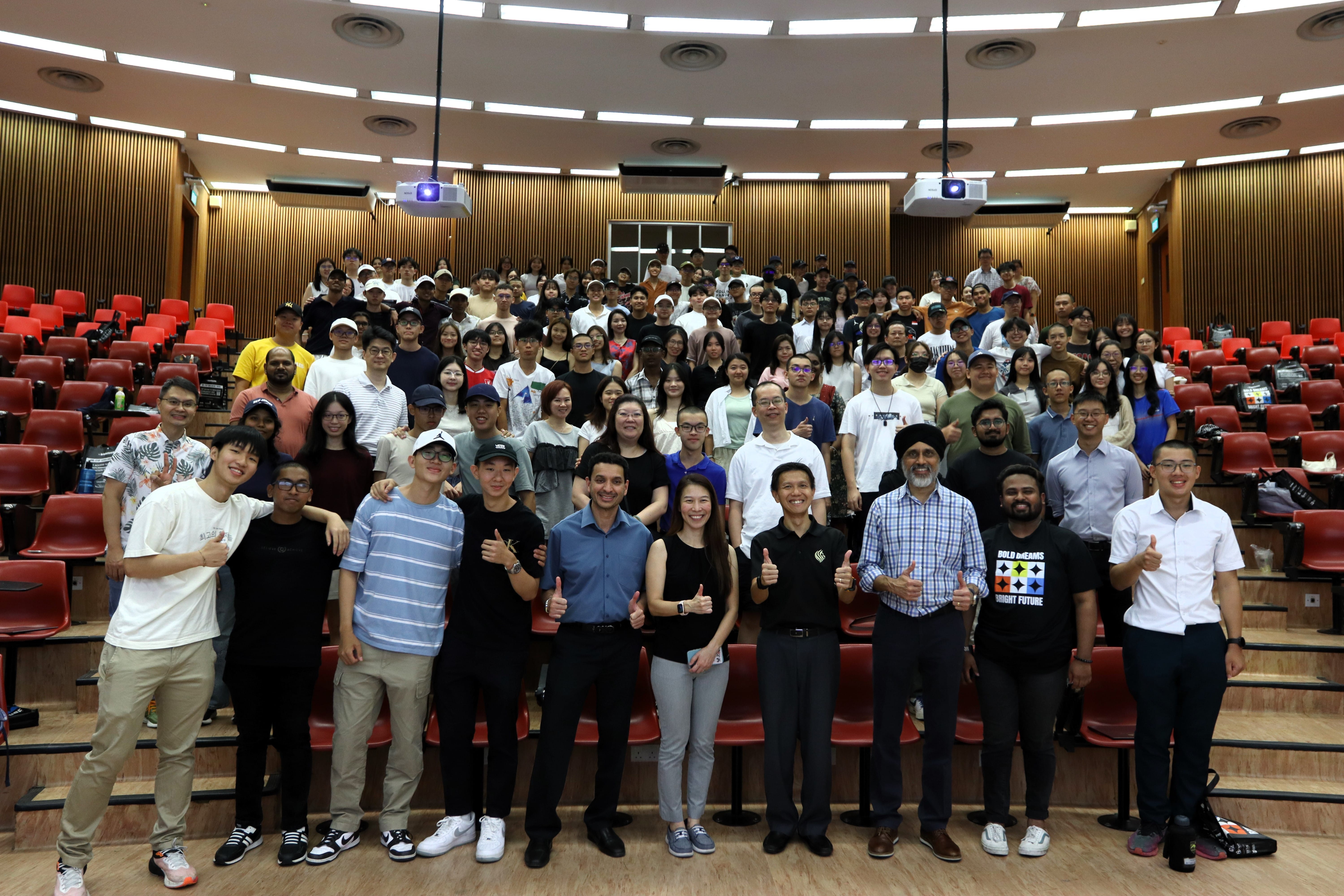 Large group posing in a lecture hall with red seats, two ceiling projectors, and several people giving thumbs up.