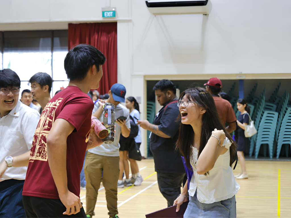 Young people laughing and socializing in an indoor gymnasium with stacked chairs and a red curtain in the background.