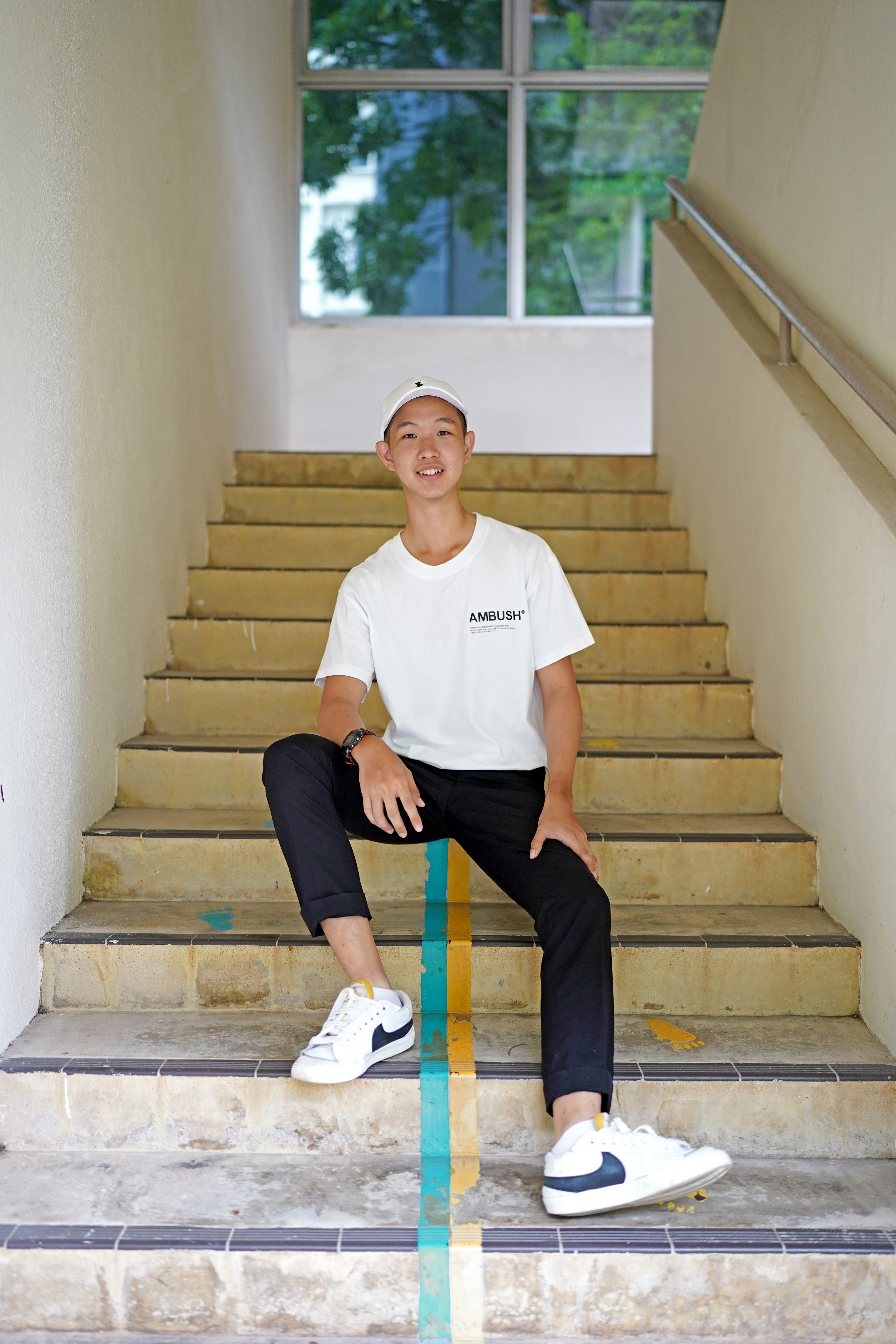 Young man in white cap and T-shirt sits on worn stairs with a blue-yellow stripe and window behind.