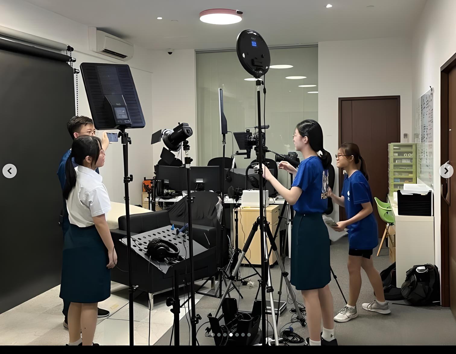Four students operate cameras and studio lights in an indoor filming setup with tripods and lighting equipment.