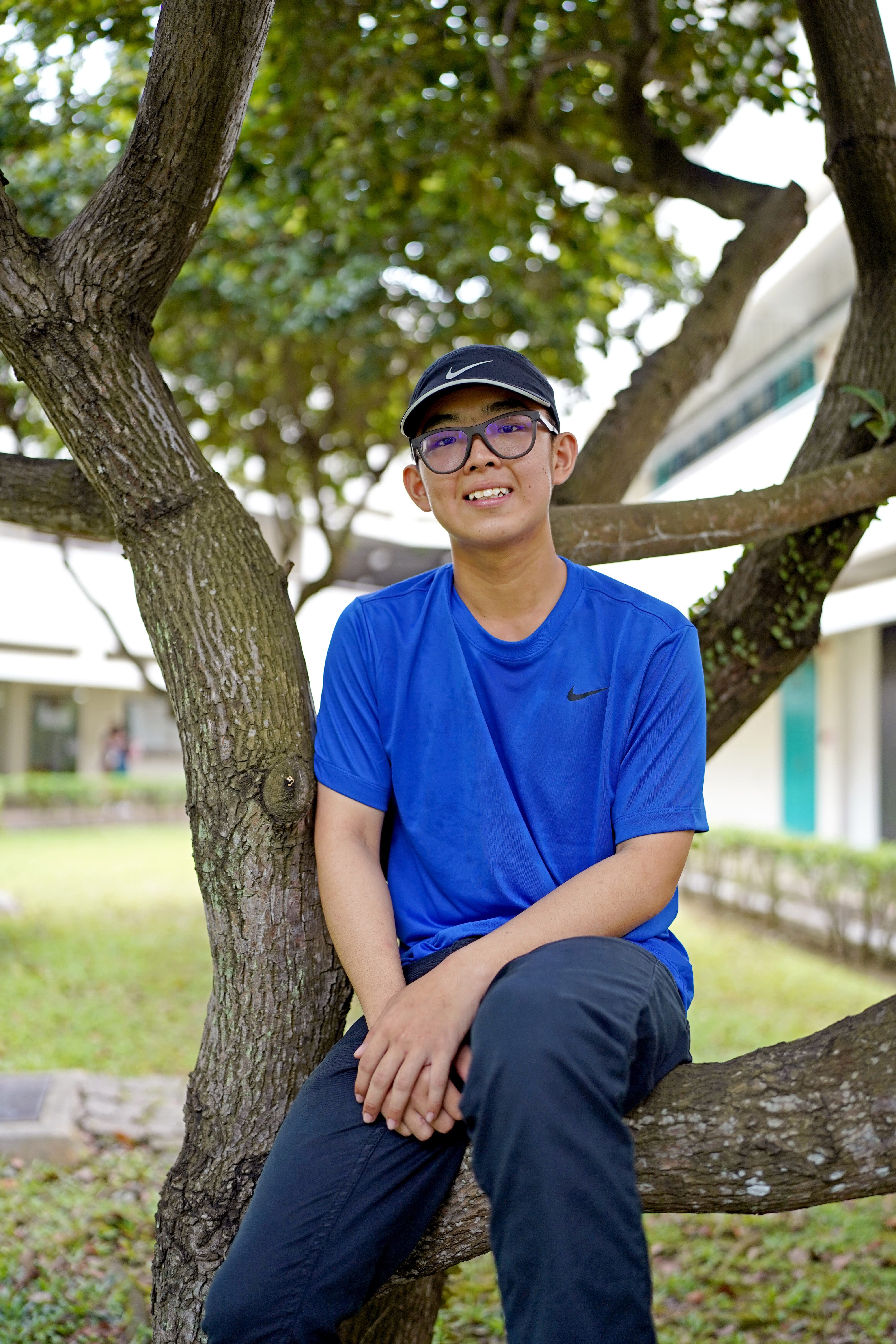Young man in blue shirt and black cap sits on a tree branch, smiling, with buildings behind.