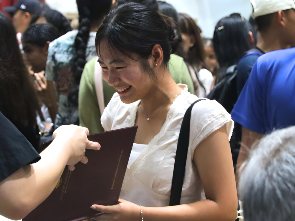 A smiling A-Level graduate collects her A-Level certificate in a crowded indoor setting.