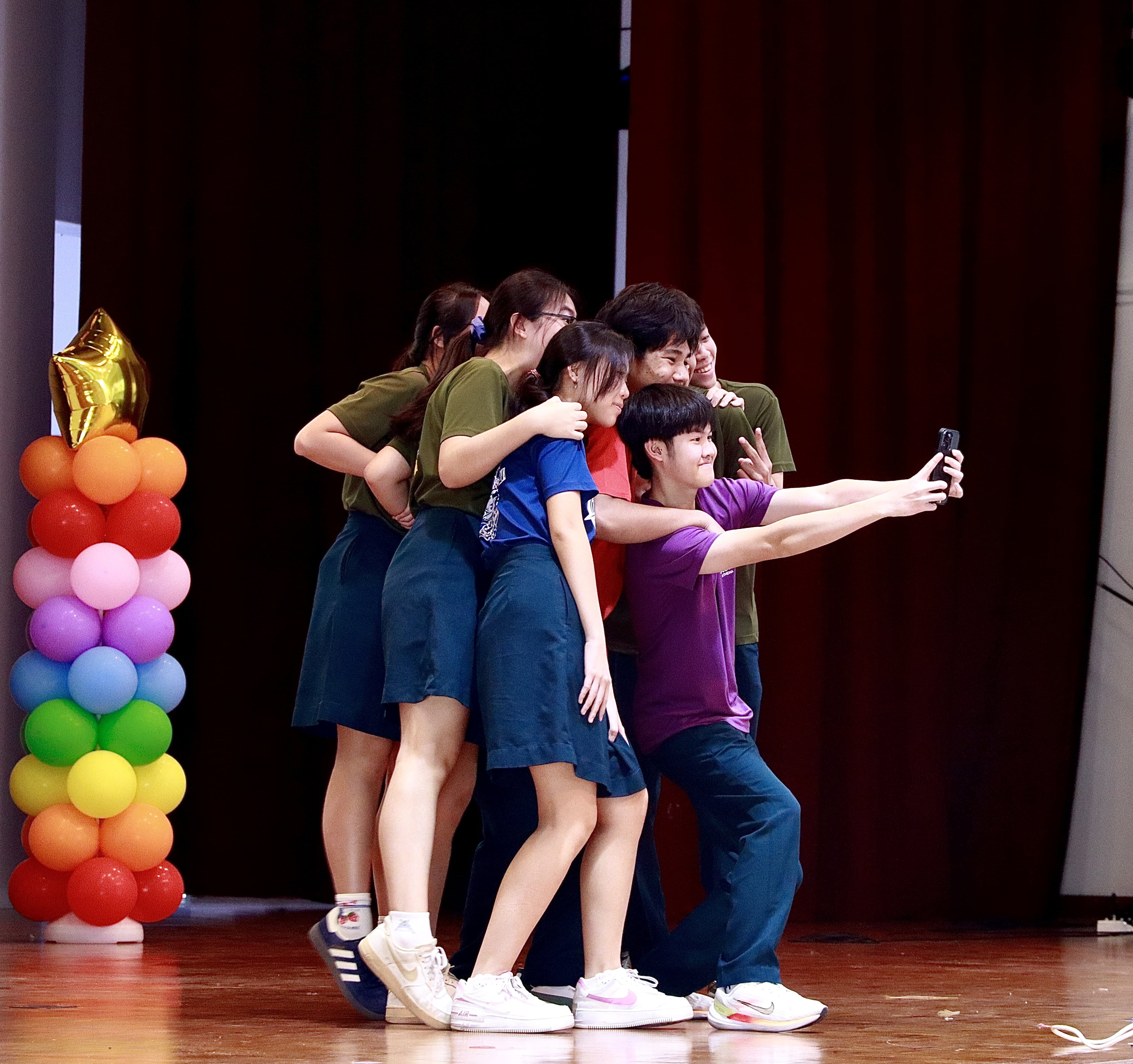 Five young people huddle together on a stage for a group selfie, with a colorful balloon tower on the left.