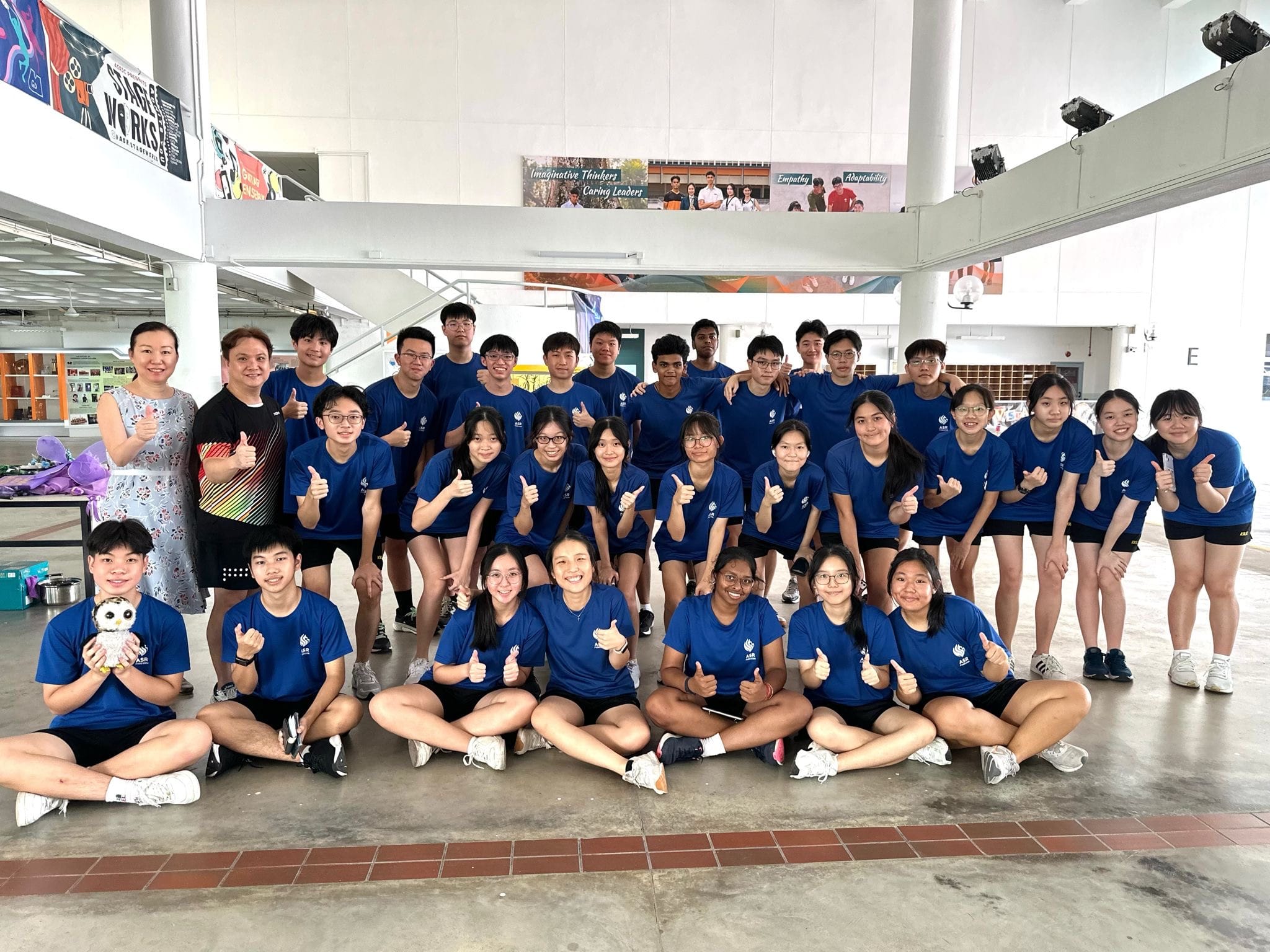 A large group of students in blue shirts giving thumbs up, posing with two teachers inside a school building.
