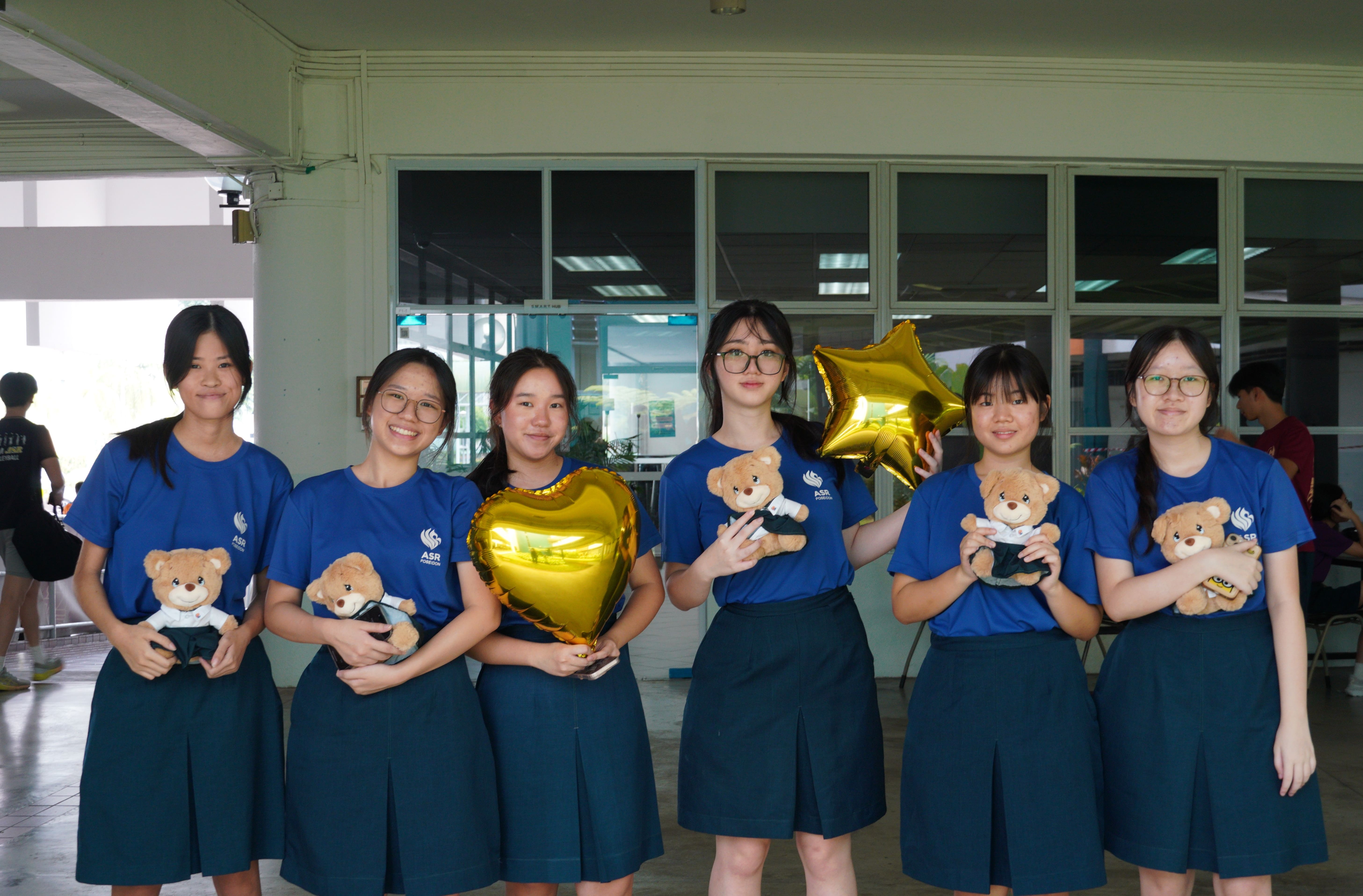 Six girls in blue school uniforms pose together holding teddy bears and gold foil balloons shaped like a heart and star.