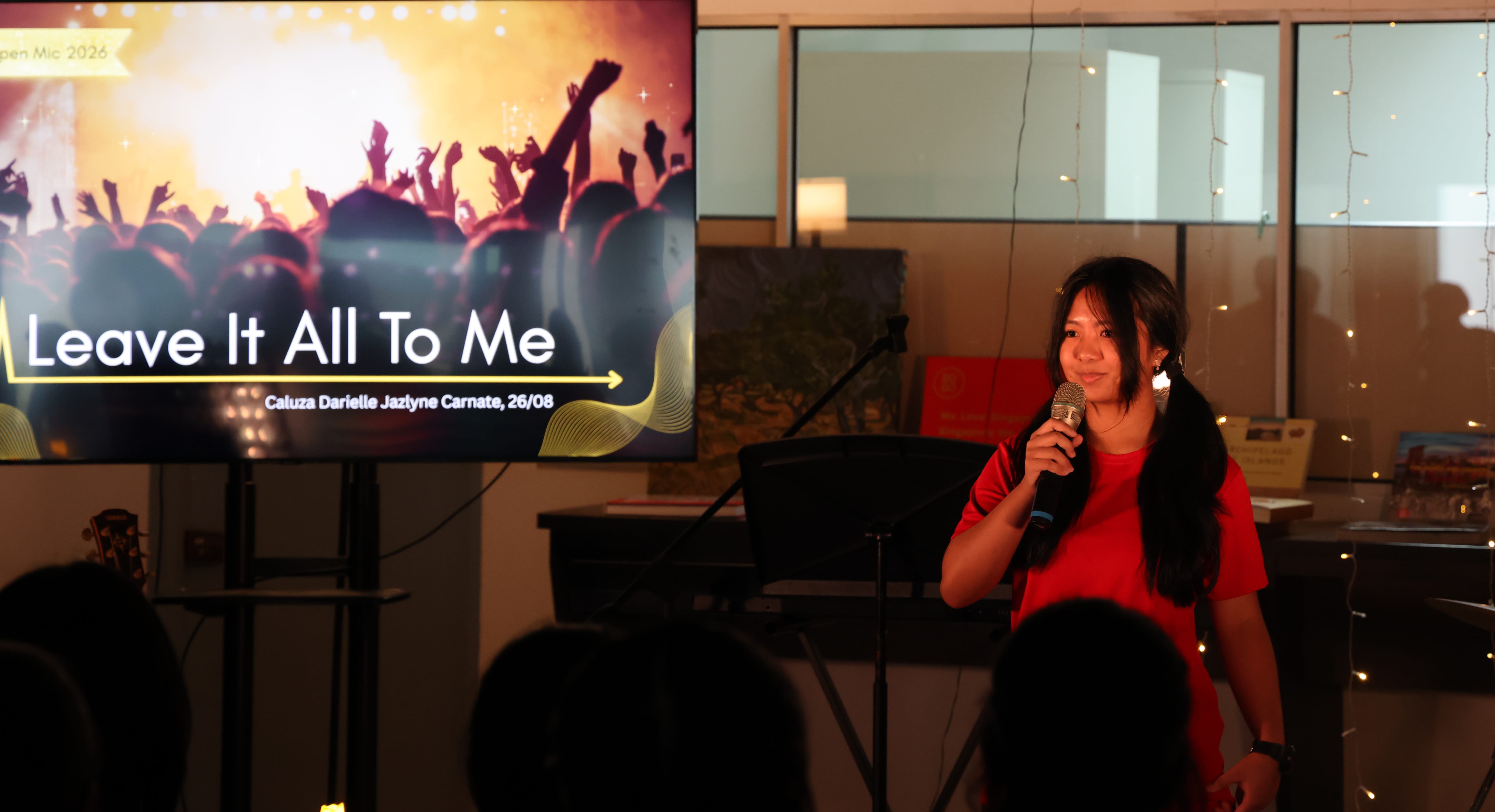 Woman in red top holds a microphone at an open mic event, with a screen displaying "Leave It All To Me" behind her.