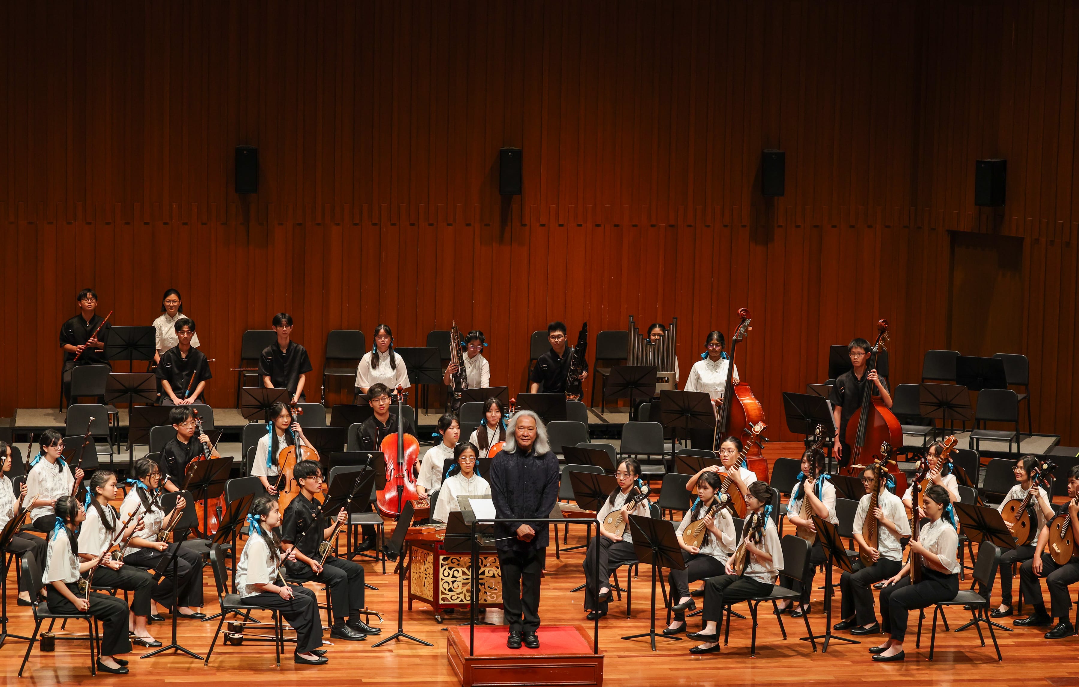 A conductor stands on a podium before a large youth Chinese orchestra on a wooden concert stage.