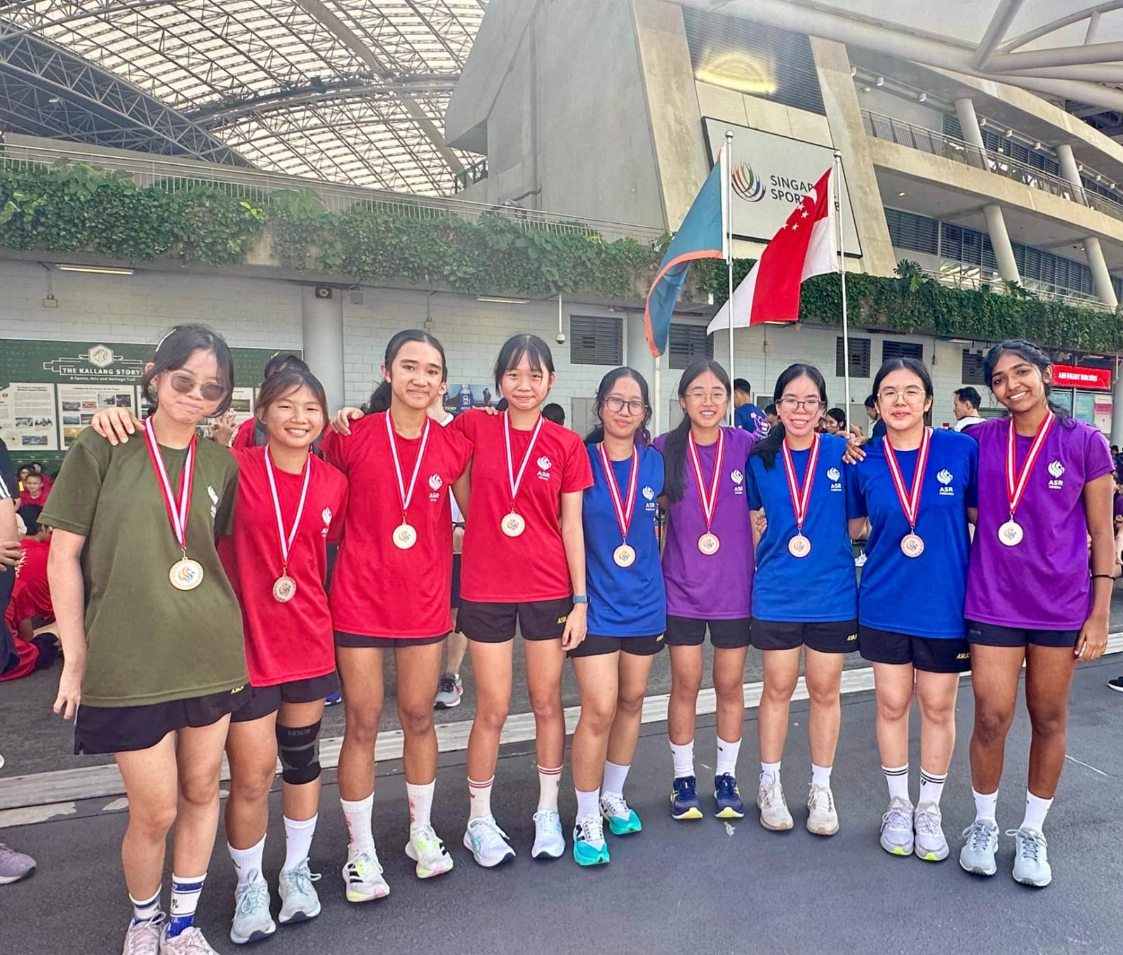 Ten young women wearing red, blue, and purple shirts with medals pose together outside Singapore Sports Hub.