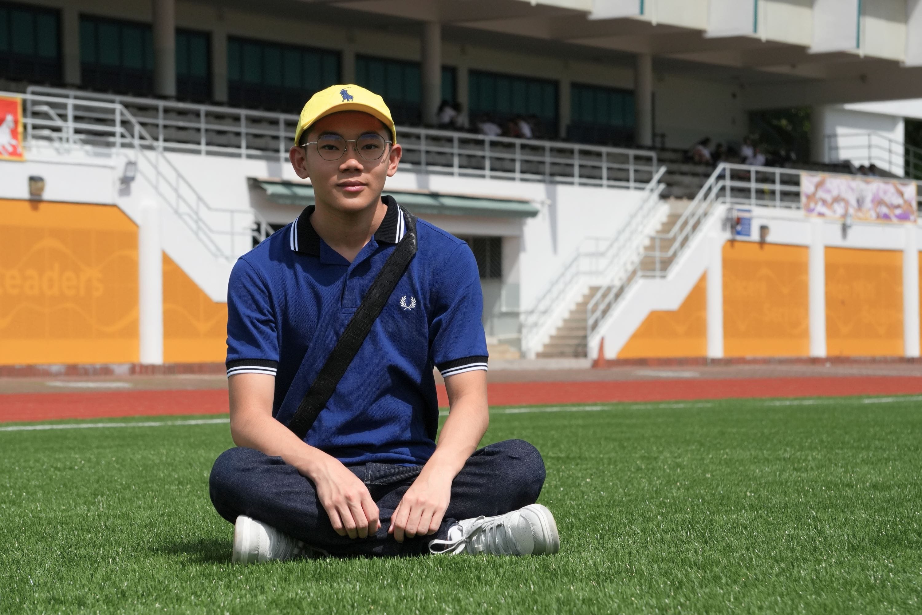 Young man in yellow cap, glasses, and blue polo sits cross-legged on green grass at a stadium.