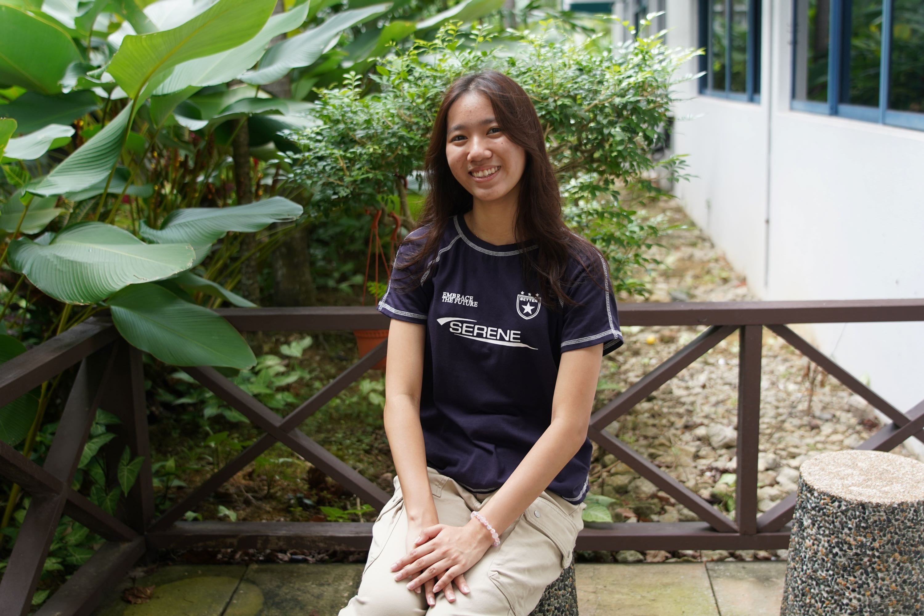 Young woman with long dark hair smiling, wearing a navy Serene jersey, seated outdoors near a wooden railing and greenery.