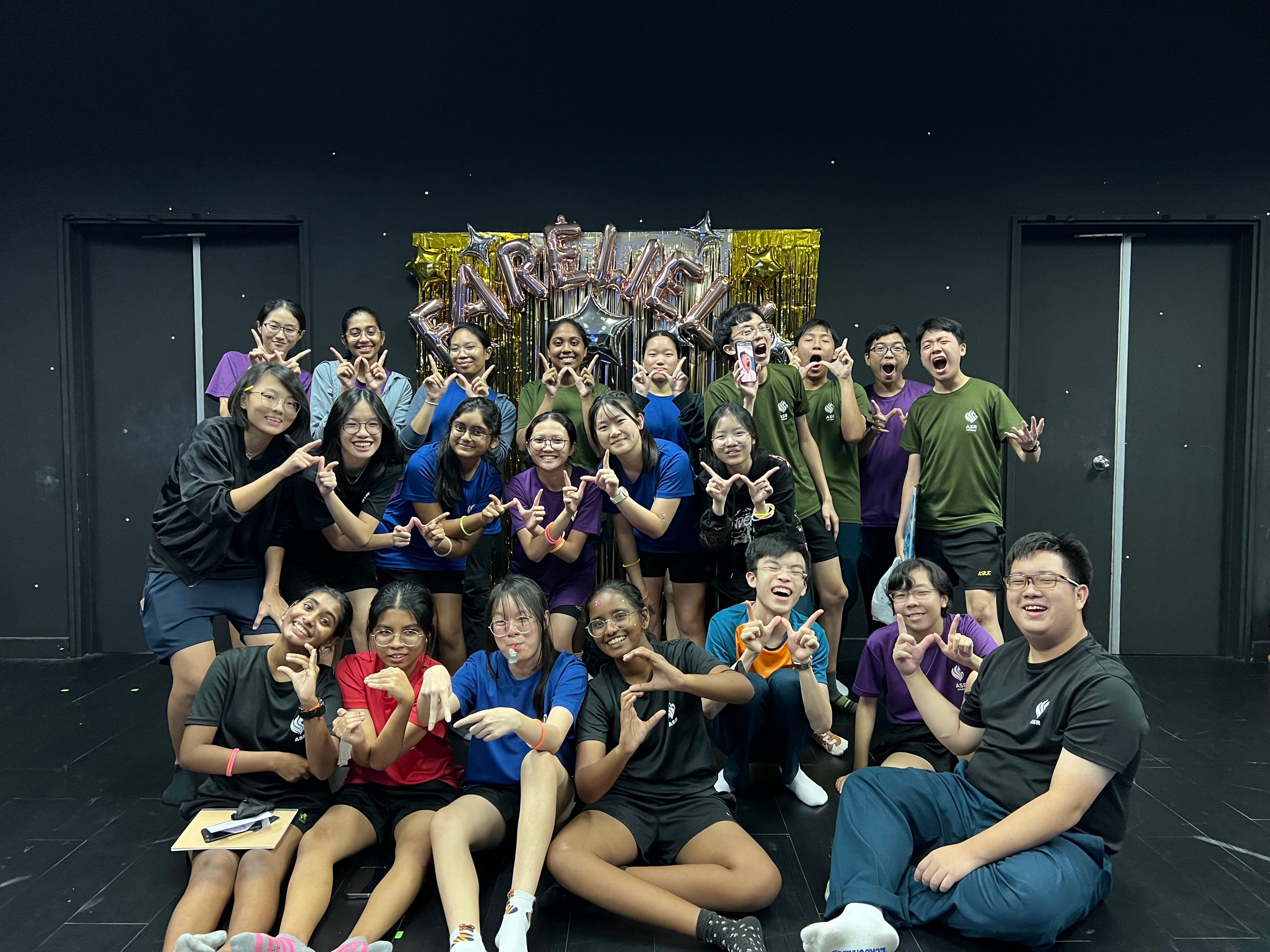 A large group of smiling teens pose with peace signs in front of a Farewell balloon banner with gold fringe.