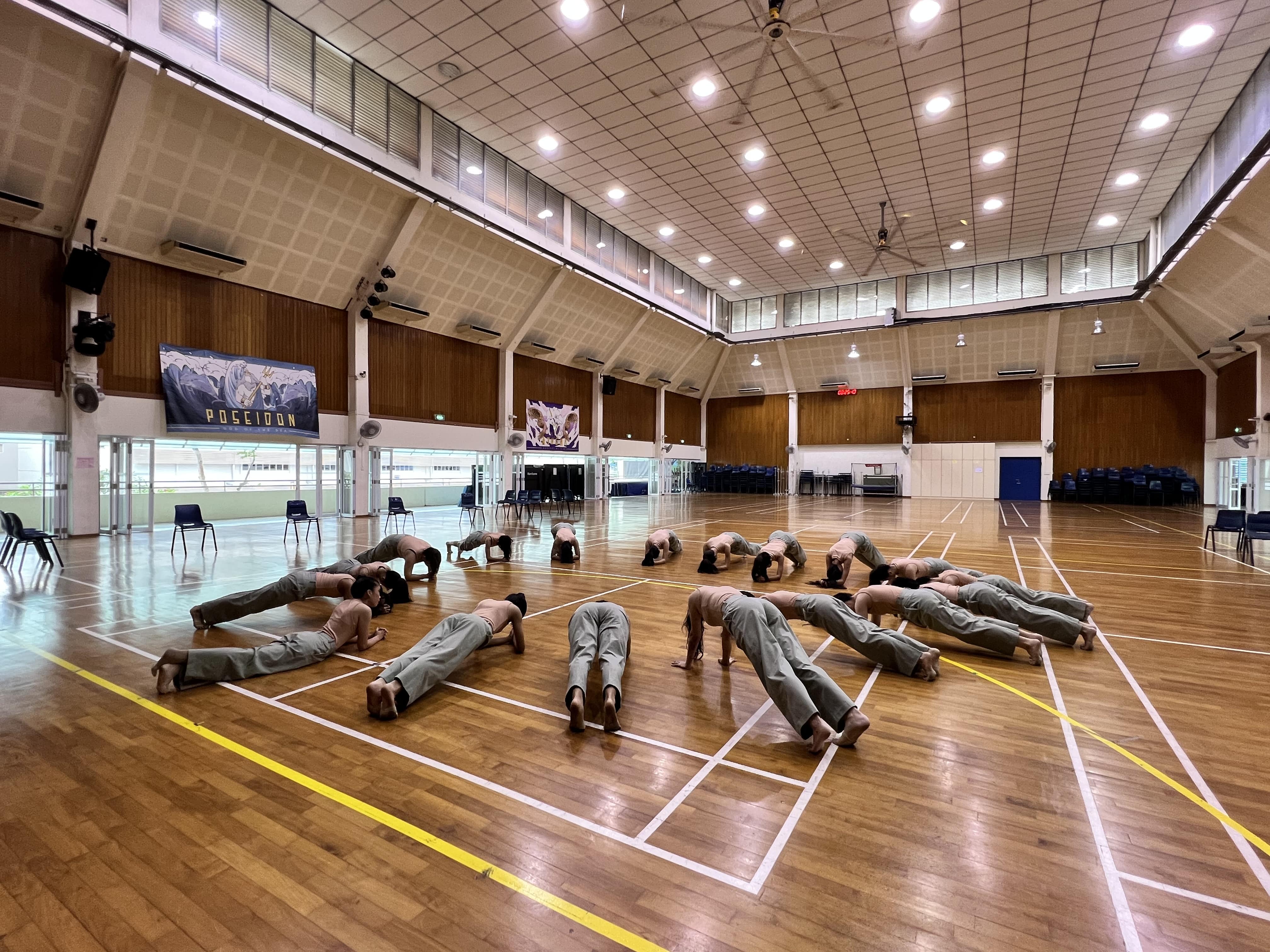 A group of performers in grey pants rehearse synchronized floor movements in a large indoor hall with a wooden floor.