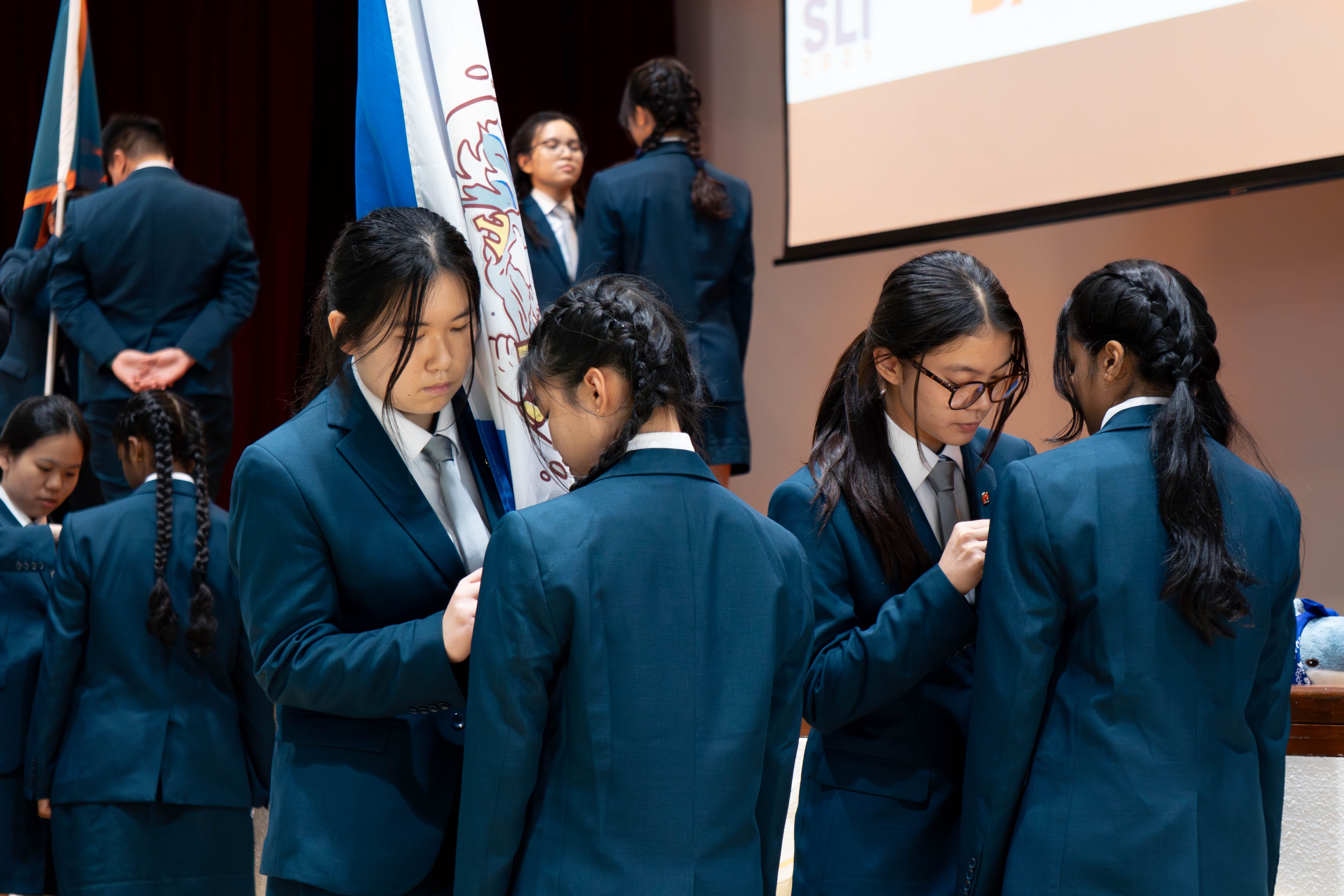 Students in teal blazers and ties pin badges on each other during a school ceremony, with flags and a screen visible behind them.