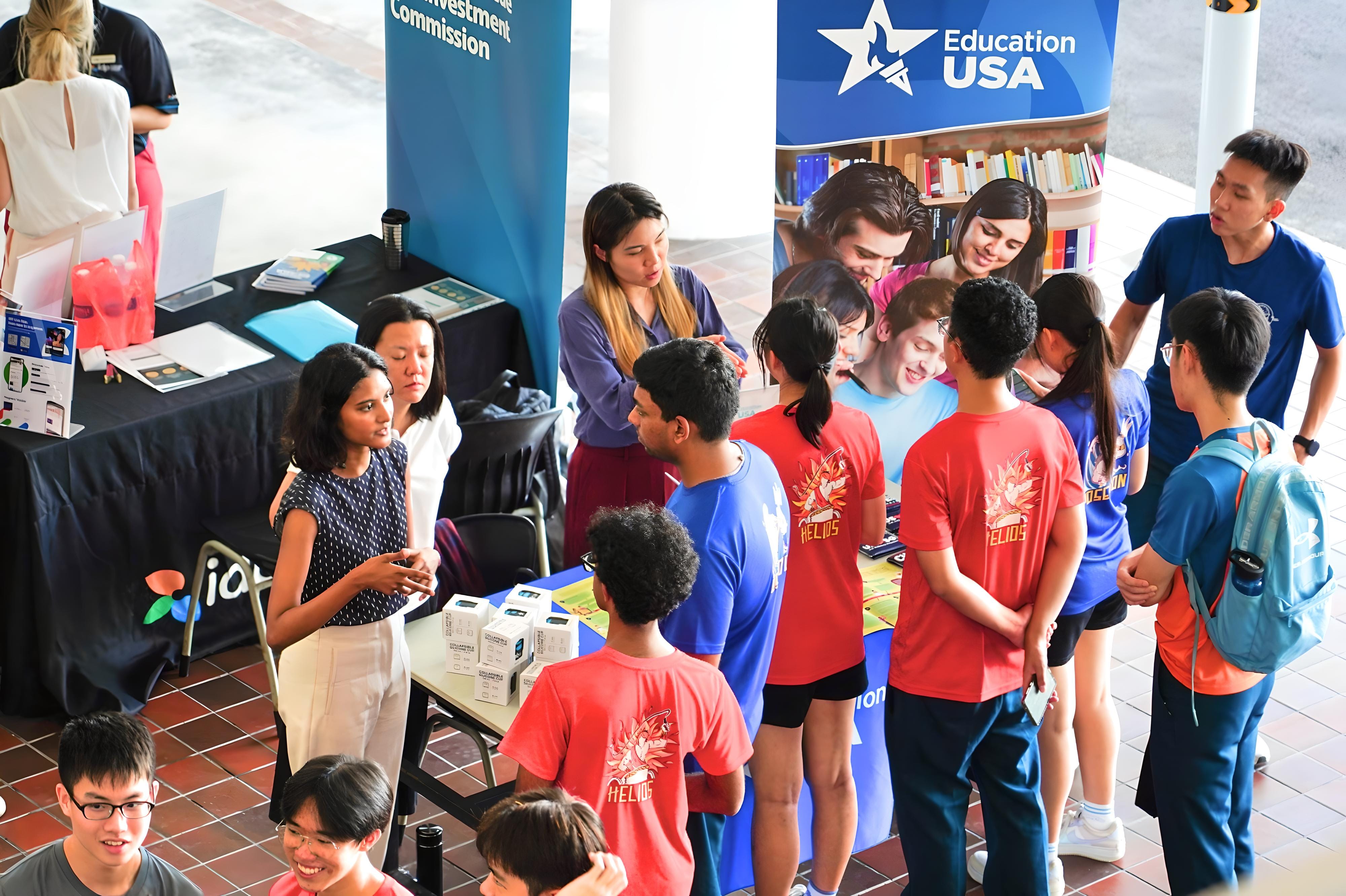 Students gather at an EducationUSA booth, talking with staff and viewing materials at an indoor fair.