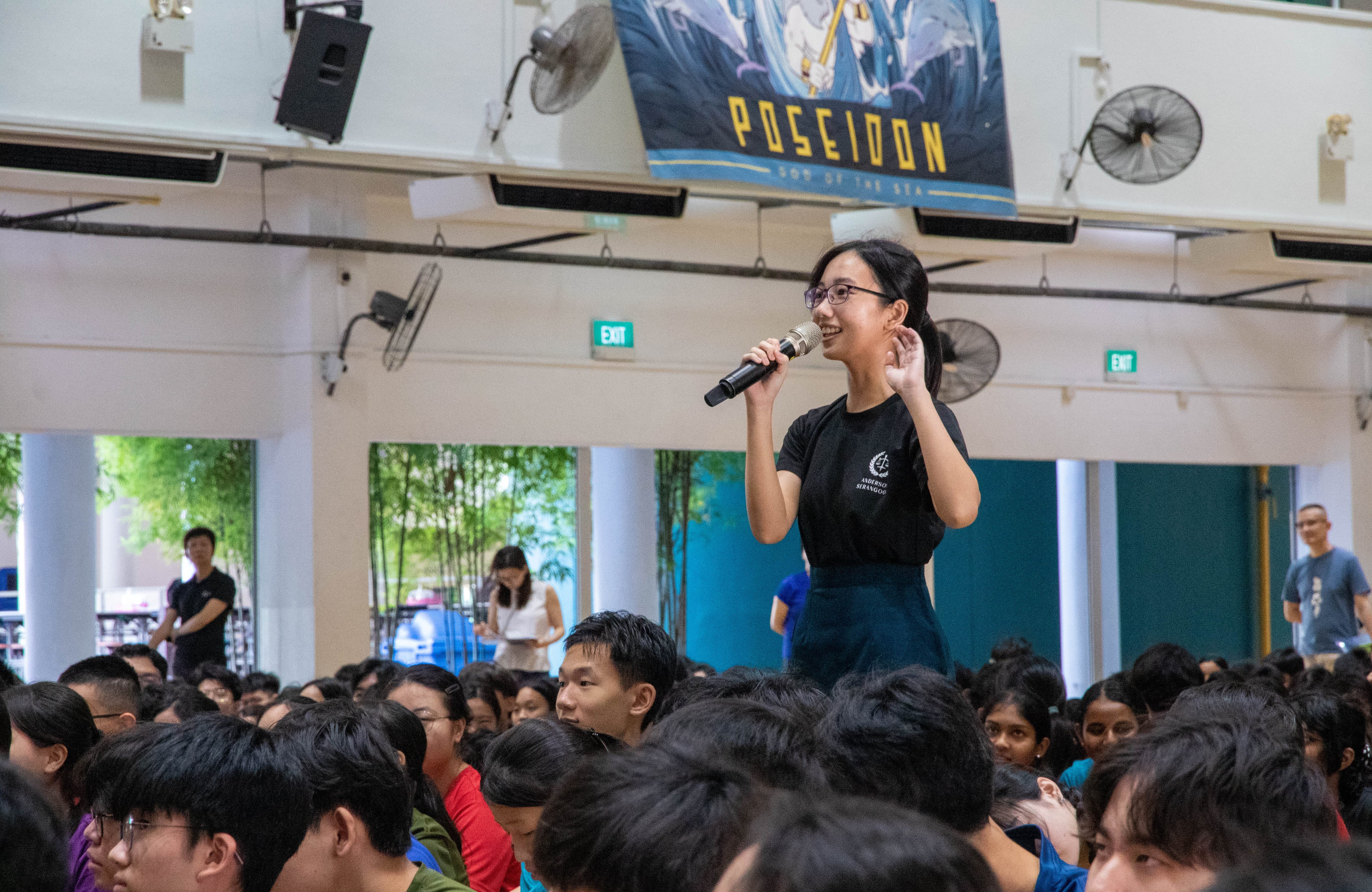 Woman with glasses speaks into a microphone while standing above a seated crowd in a hall with fans and a banner.