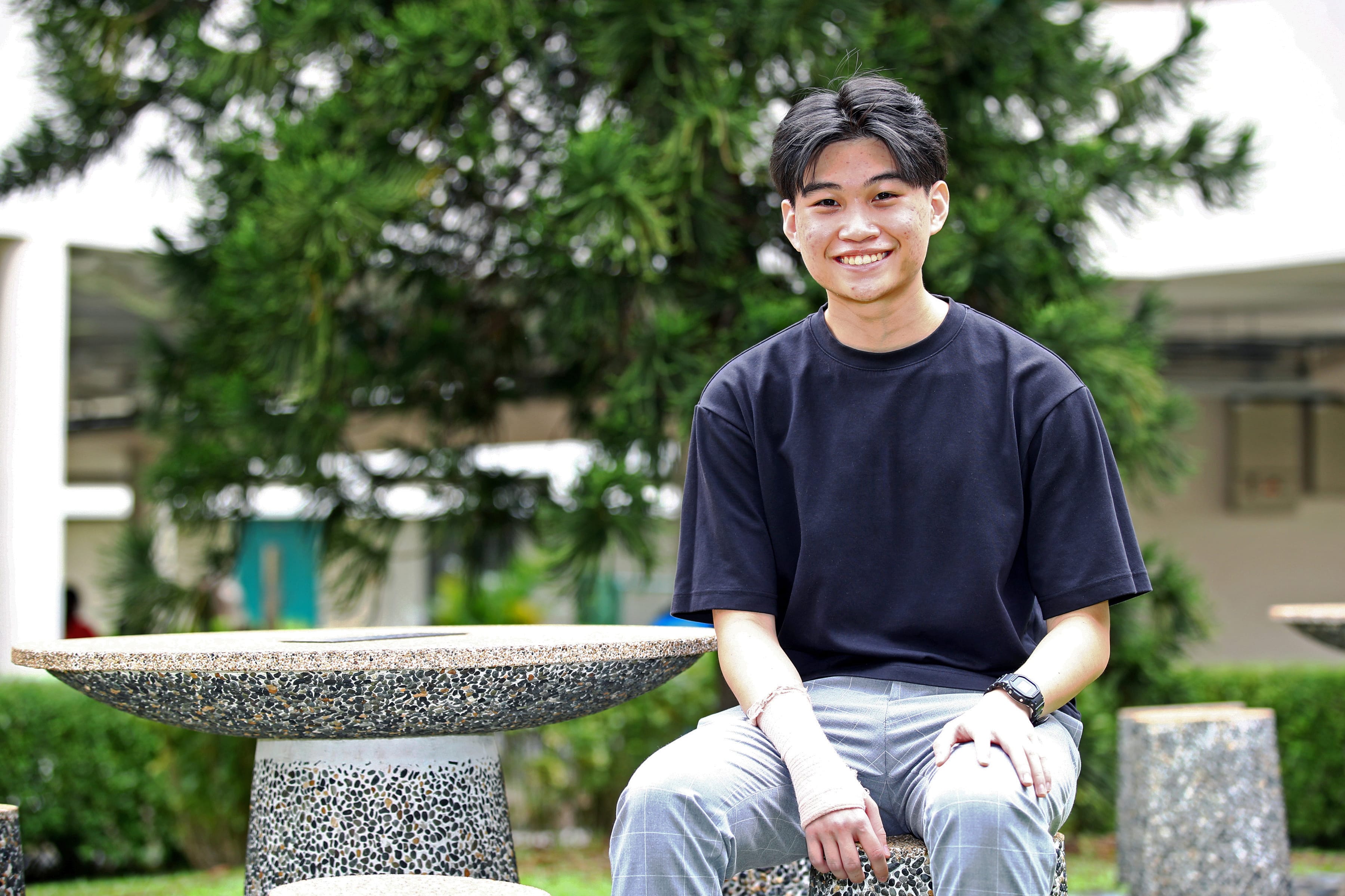 Young man in a black t-shirt smiling while seated outdoors near a stone table with trees and a building behind him.