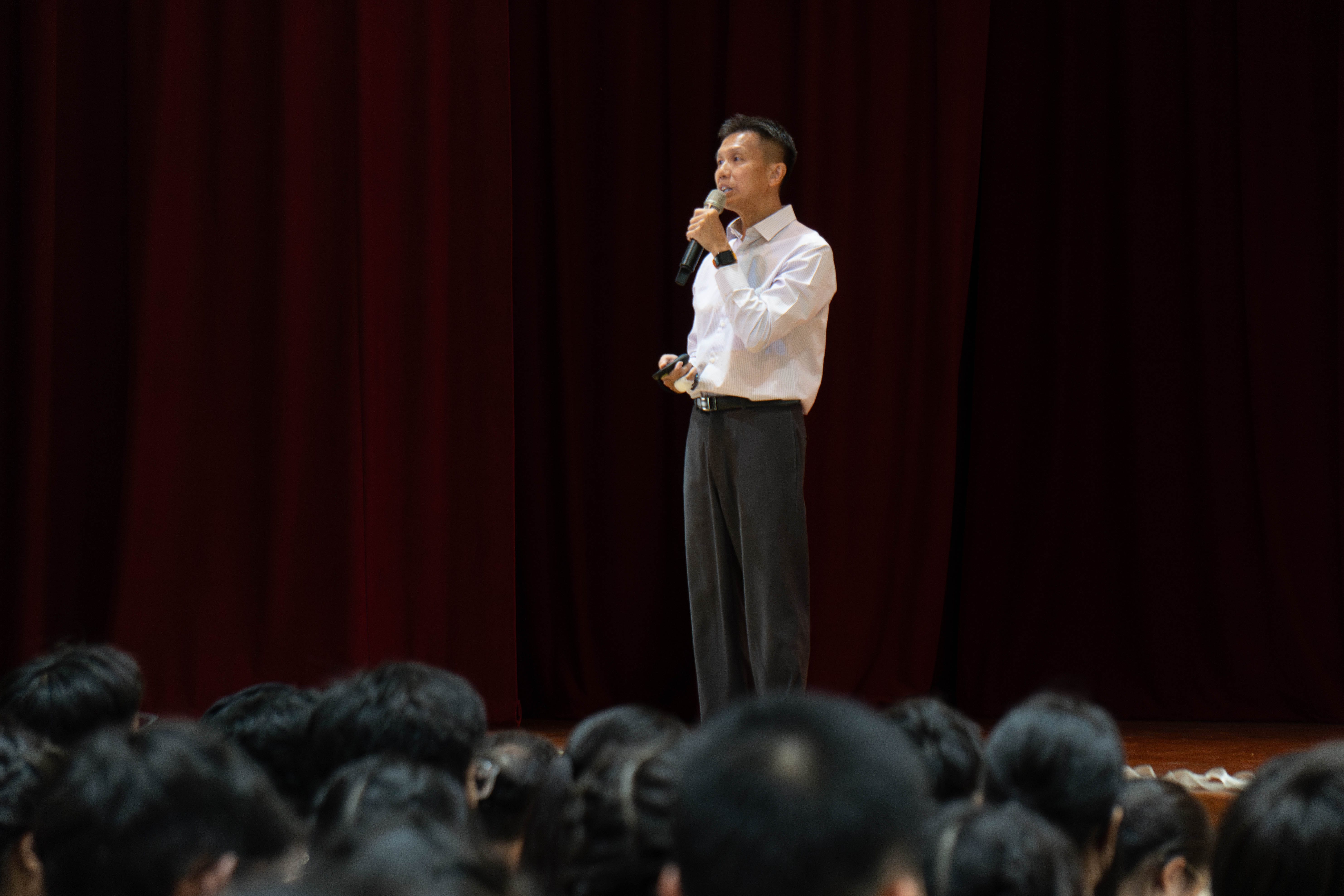 A man in a white shirt speaks into a microphone on stage before a seated audience with a red curtain behind him.