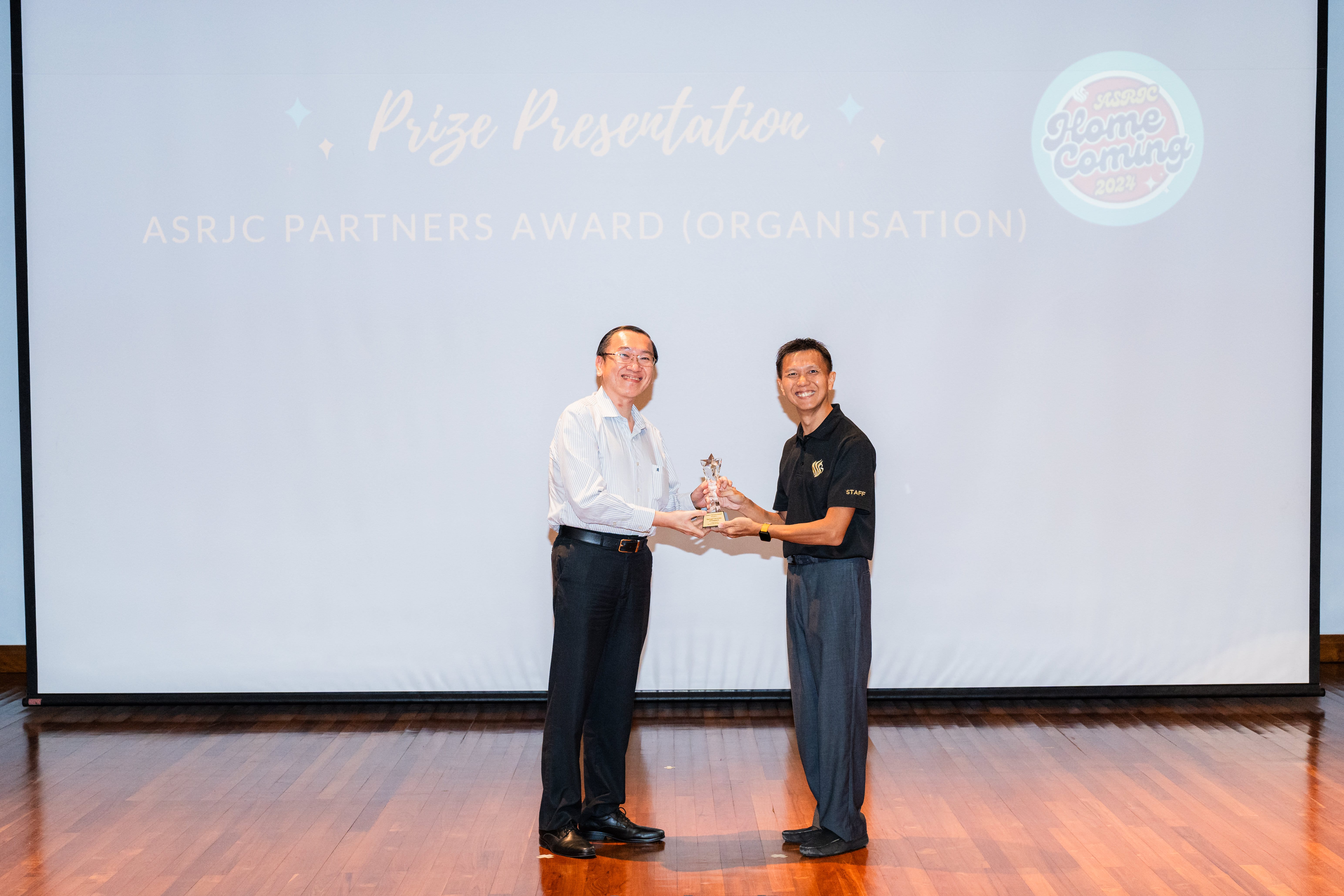 Two men smile while holding a trophy together on stage at the ASRJC Partners Award Organisation prize presentation.
