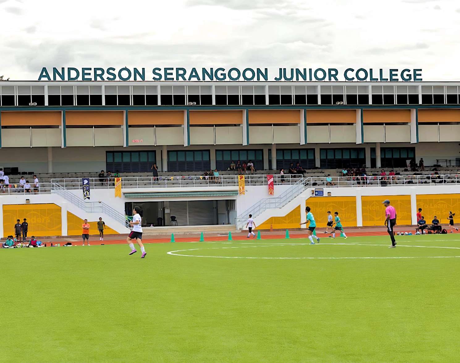Students play football on a green field in front of Anderson Serangoon Junior College's multi-story building.