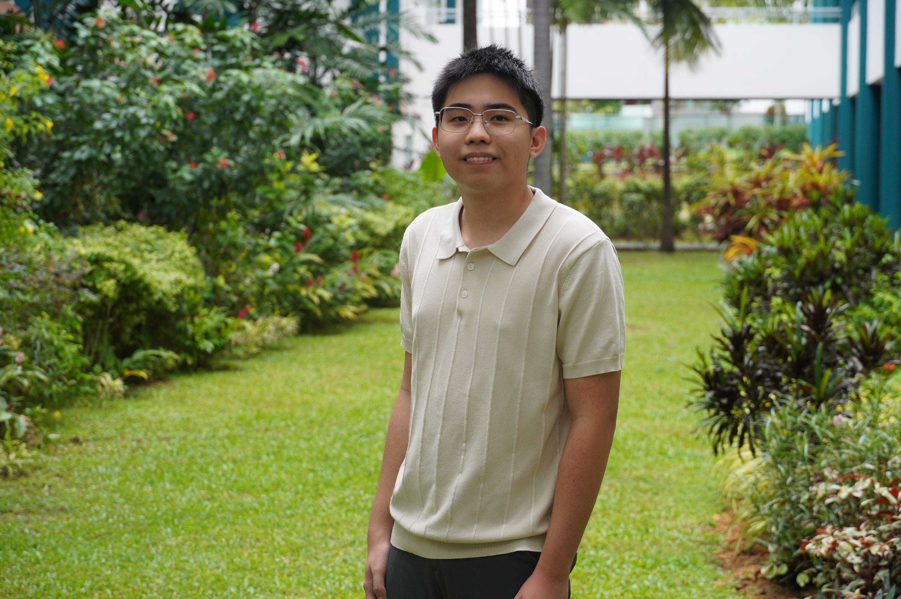 Young man with glasses wearing a cream polo shirt, smiling outdoors in a lush green garden with tropical plants.