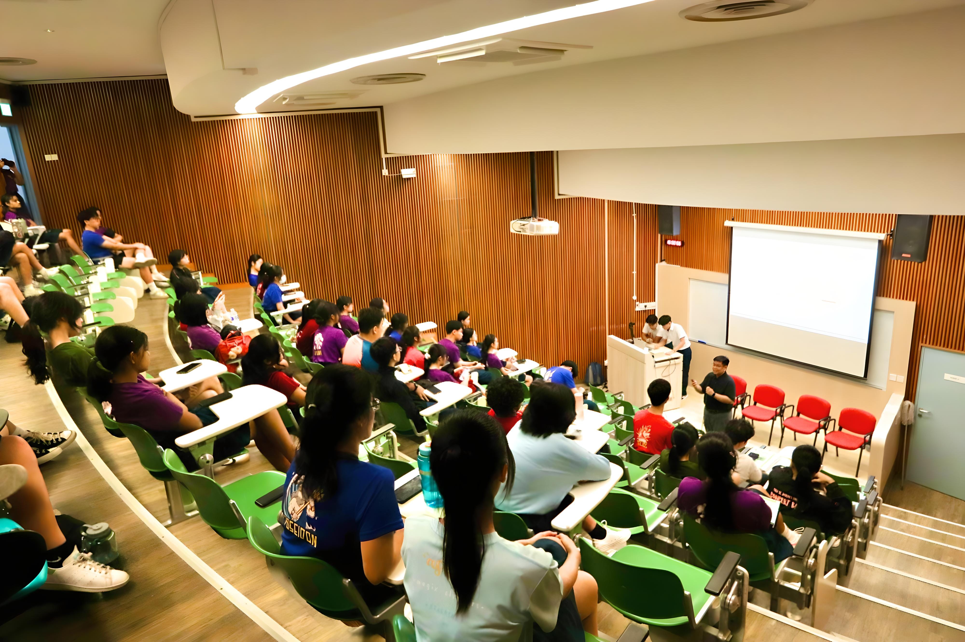 Students seated in green chairs in a lecture hall, watching a presenter beside a projection screen.