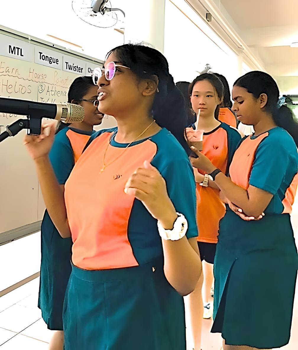 A girl in orange and teal uniform speaks into a microphone while several classmates stand behind her indoors.