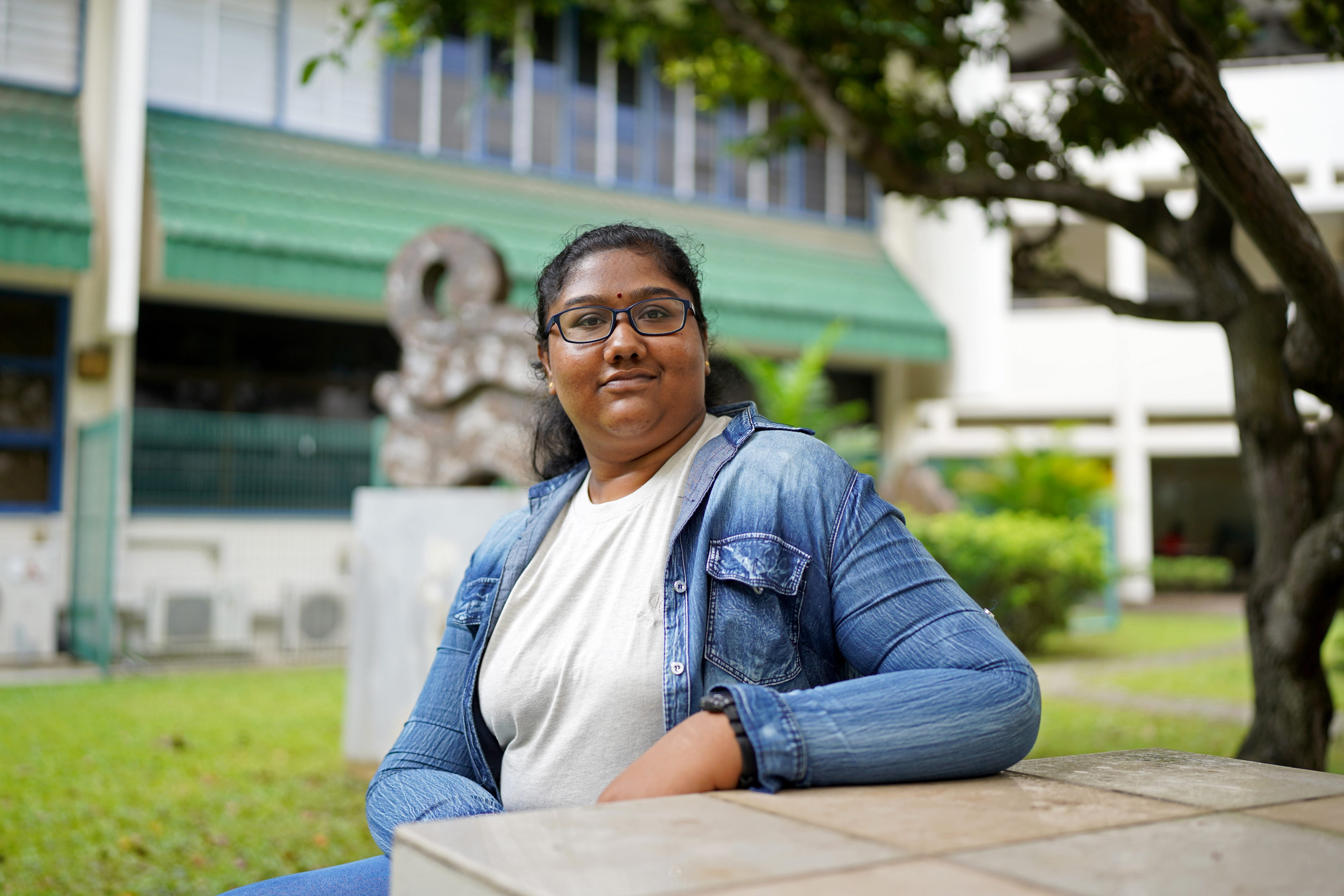 Young woman with glasses in denim jacket sits at outdoor table, building and tree in background.