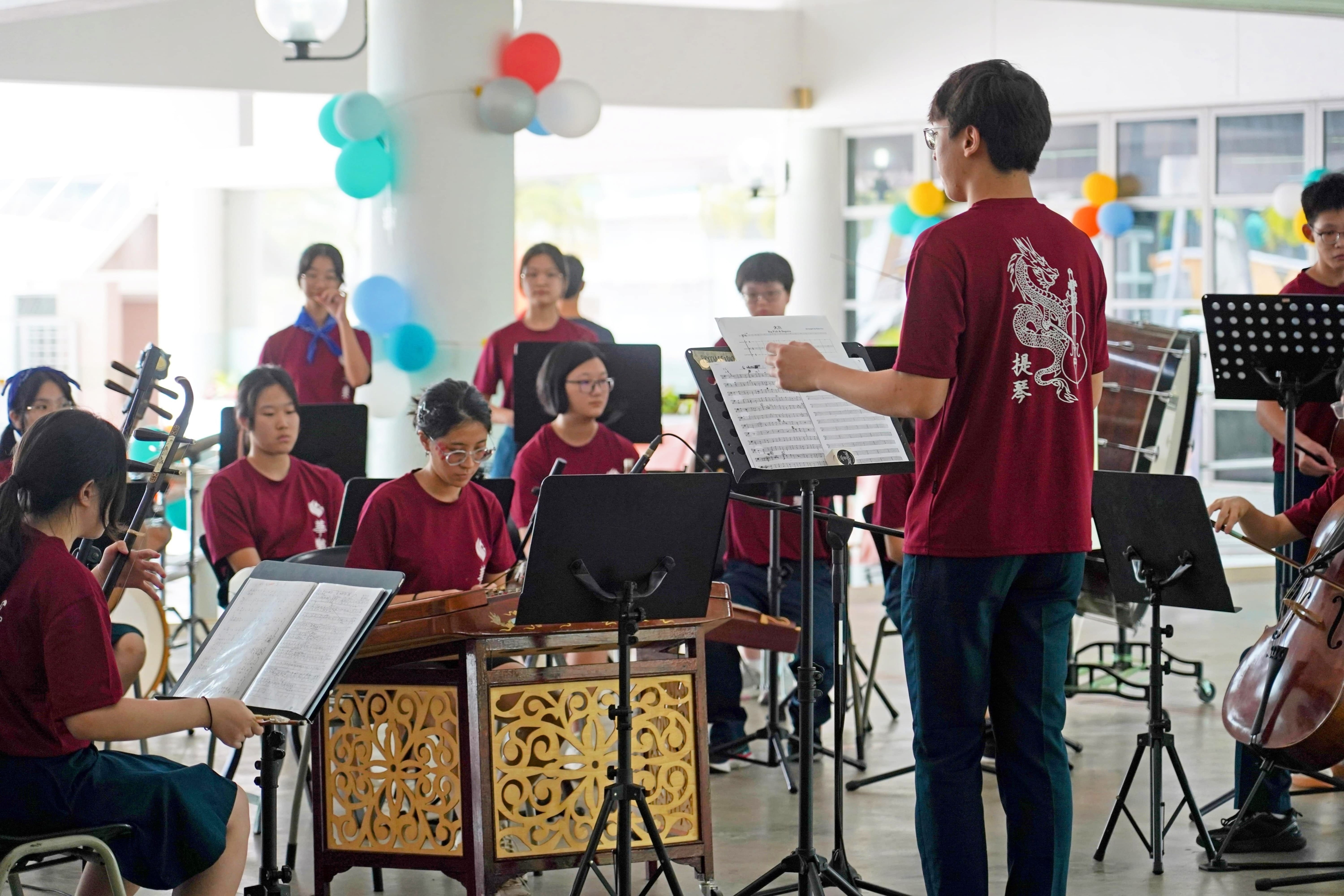 A youth Chinese orchestra in matching red shirts performs outdoors, led by a conductor holding sheet music.