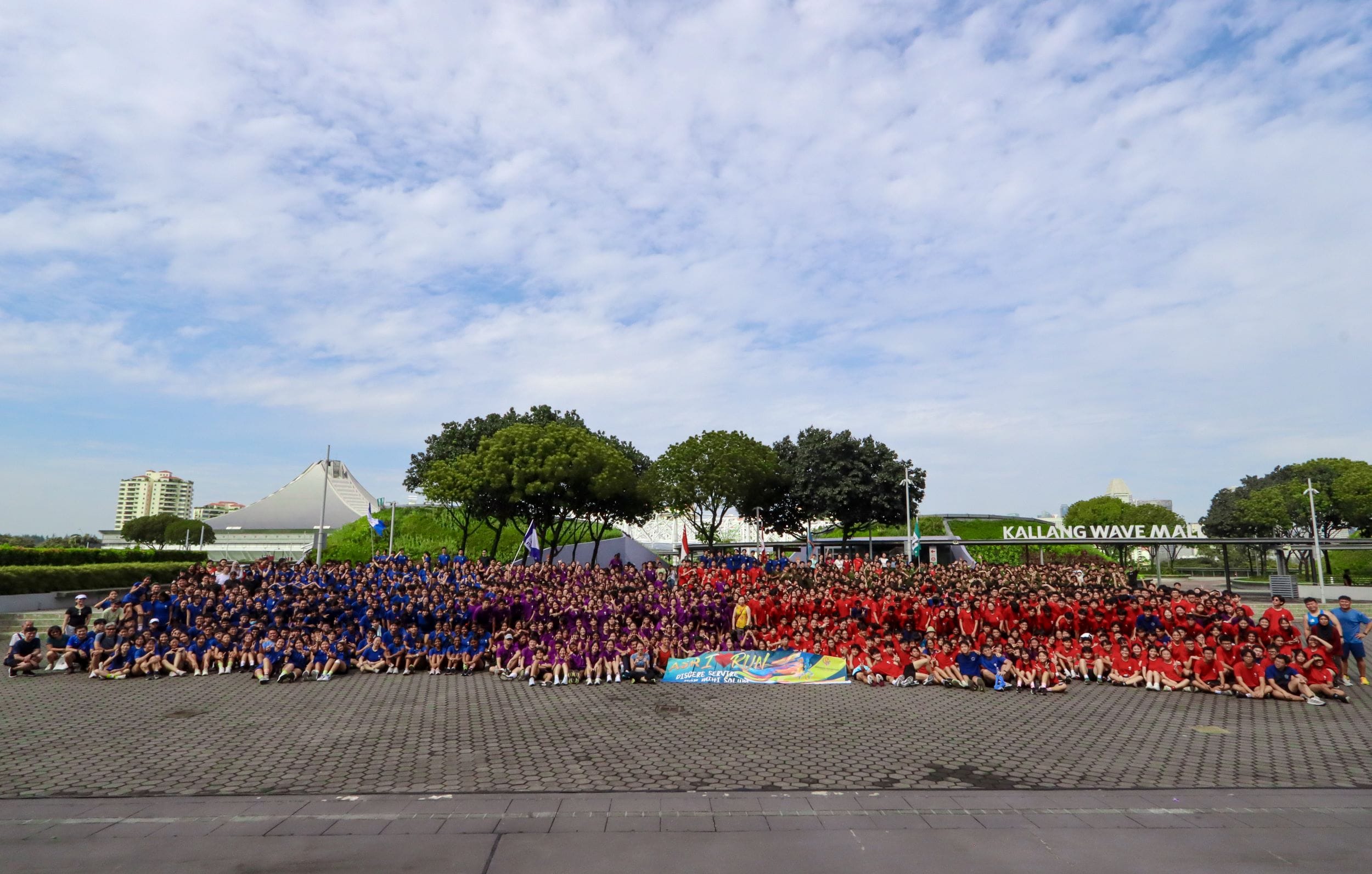 Large group photo of hundreds of people in blue, purple, and red shirts outside Kallang Wave Mall.