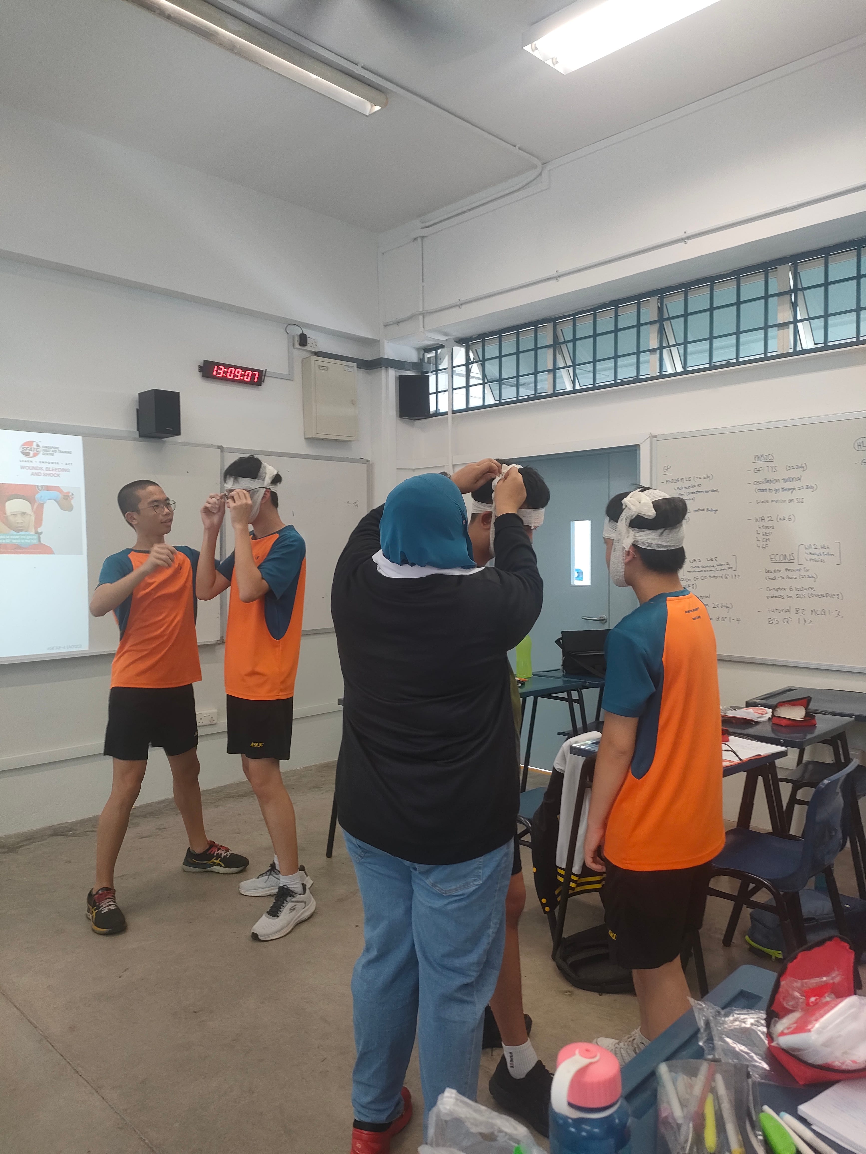 Four students in orange jerseys wear bandaged head wraps in a classroom with whiteboards and a projector.